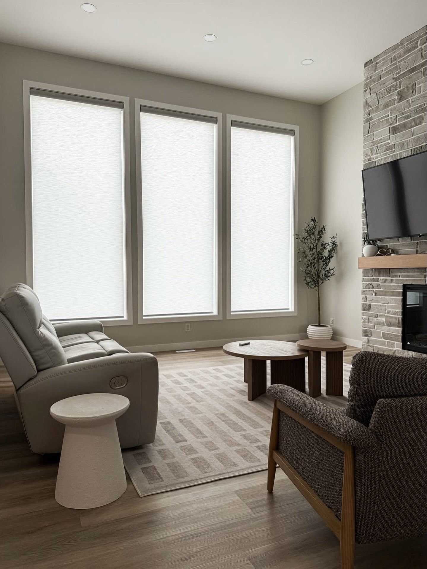 Living room with three large windows, gray sofa, small table, armchair, and stone fireplace with TV.