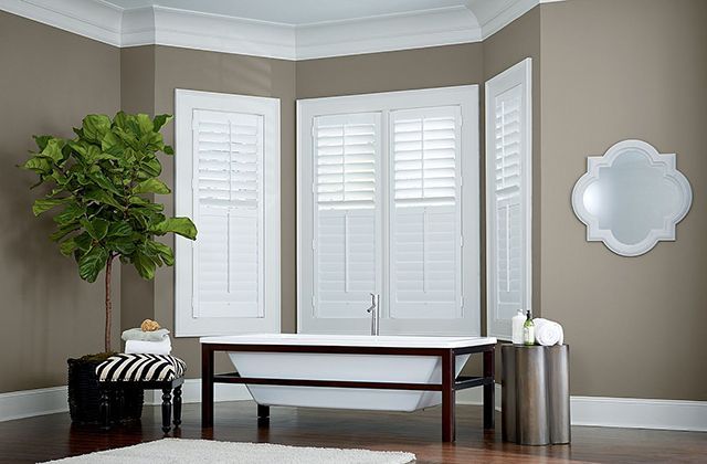 Bathroom with white shutters, freestanding tub, green plant, and decorative mirror.