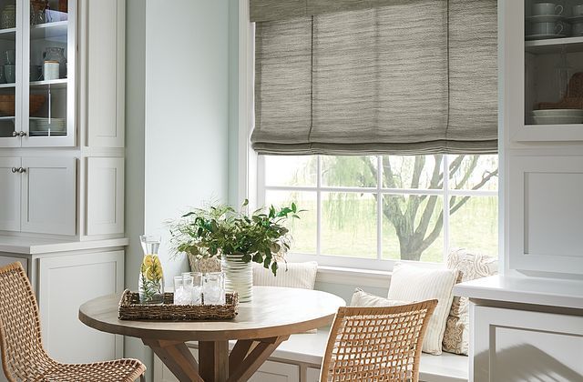 Kitchen nook with round wooden table, woven chairs, window seat, and woven shade.