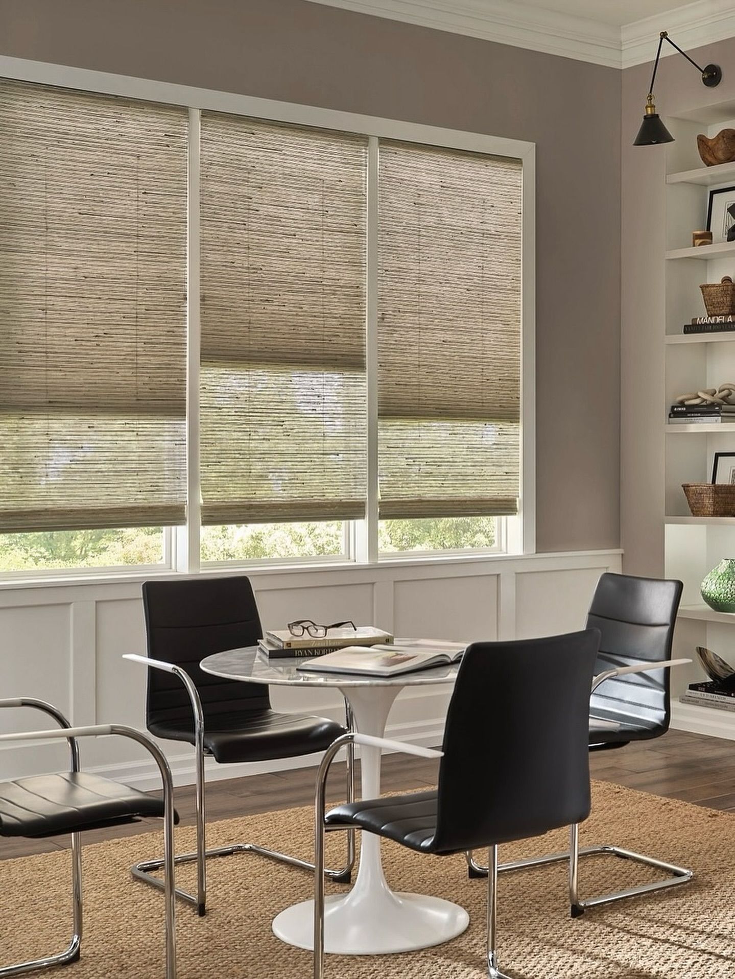 A dining room with round table and black chairs; window with blinds; shelves on wall.