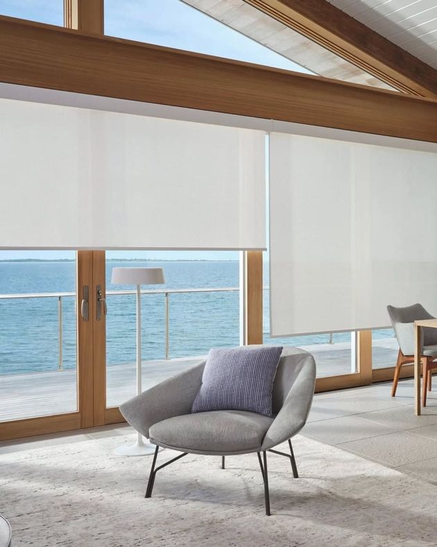 Gray armchair with blue pillow in front of large window with white blinds and a view of the ocean.
