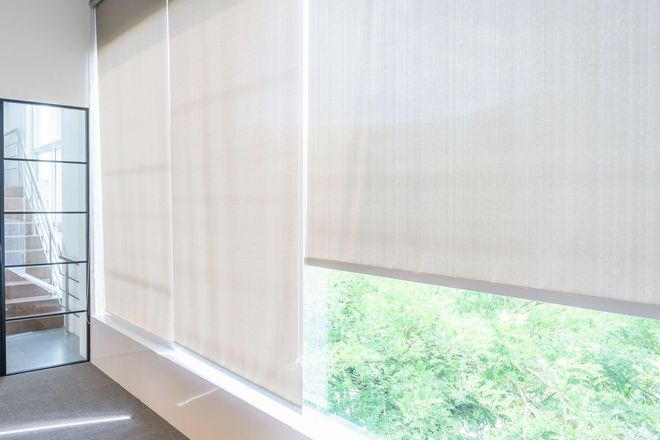 Off-white roller blinds cover large windows, with one partially raised to reveal greenery outside.