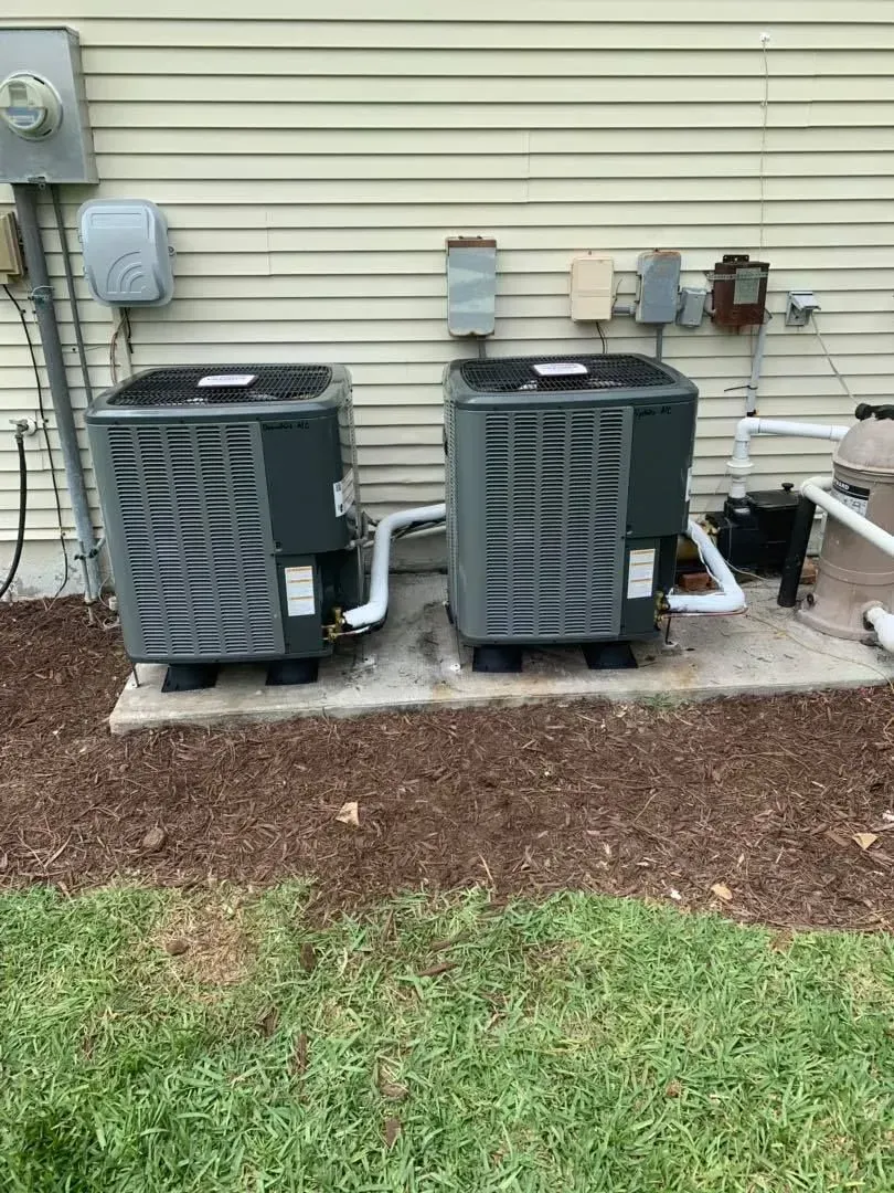 Two outdoor HVAC units beside a house with utility meters and electrical boxes on the siding