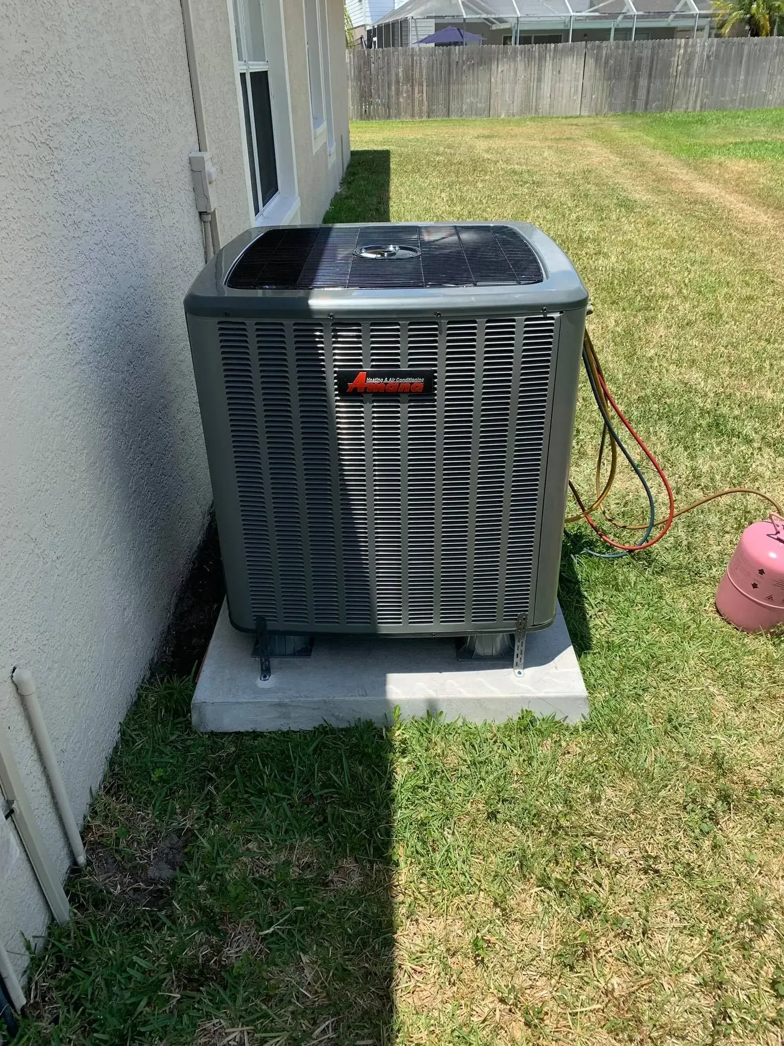 Outdoor air conditioner unit beside a house, with a red hose on the grass and sunlight casting shadows.