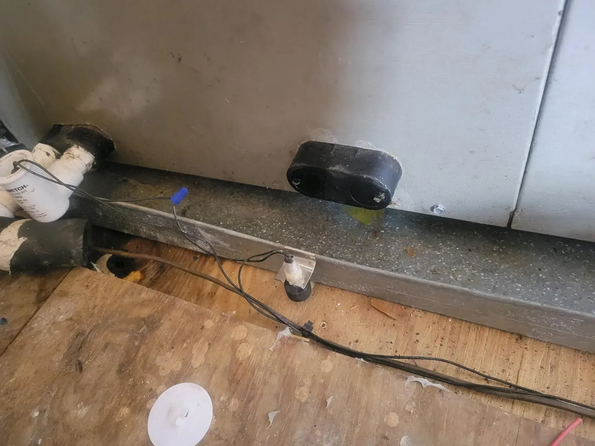 Wall base with exposed cables and a black wall plug on a dusty floor near a white container.