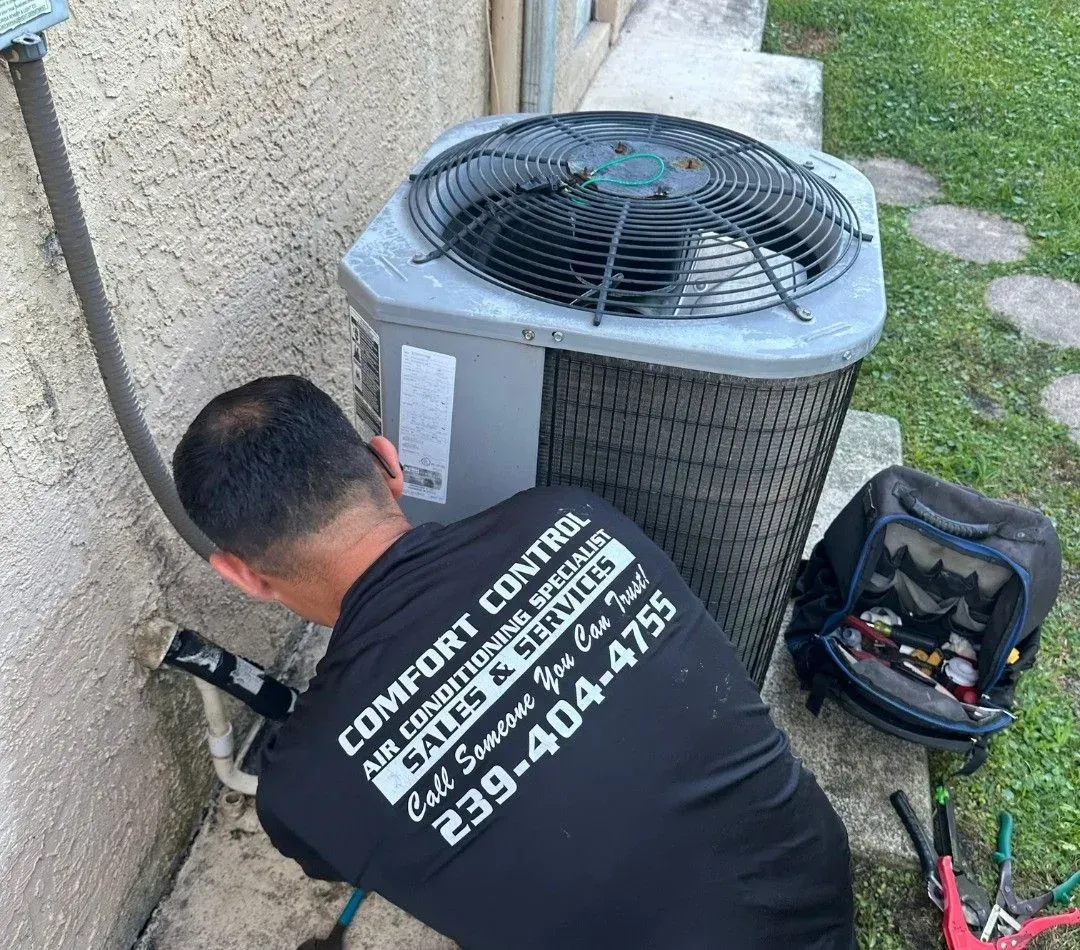 Technician working on an outdoor HVAC unit beside a house wall, with tools on the ground.