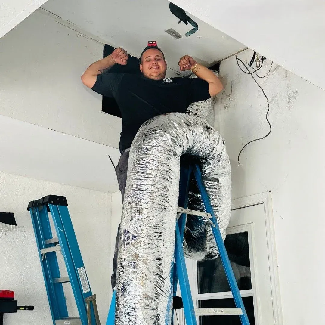 Worker standing on a ladder in a room, flexing beside large silver HVAC ducts near the ceiling