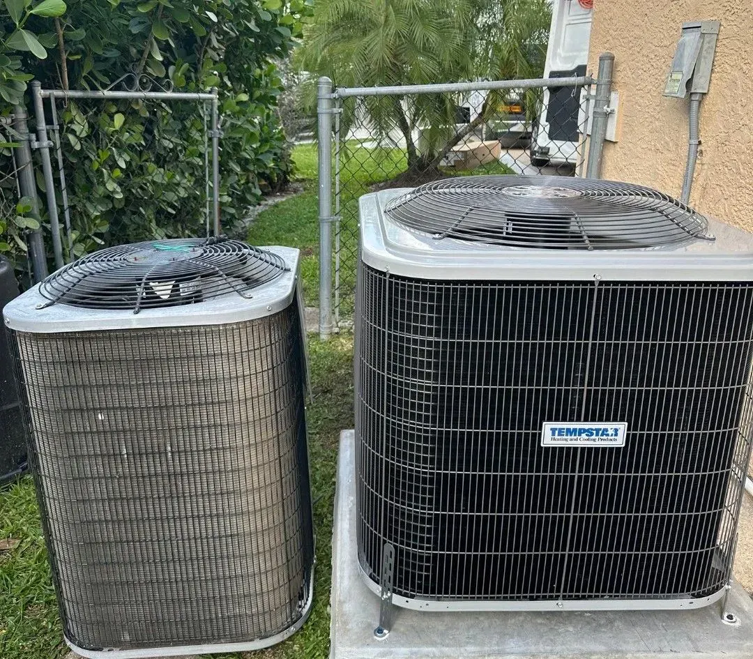 Two outdoor air conditioner condensers beside a house, one older and one newer.