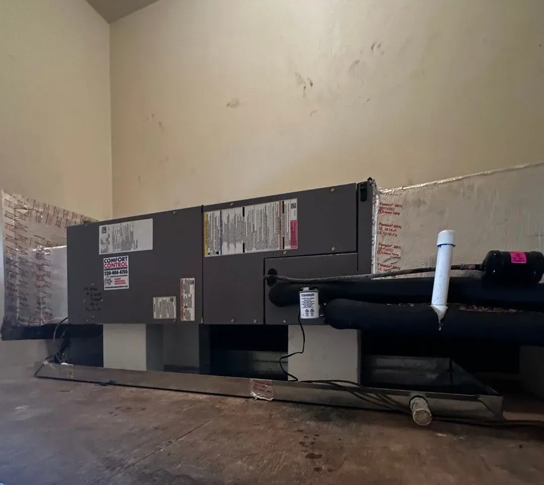 Large gray HVAC unit in a basement utility room with exposed pipes and a rough concrete floor