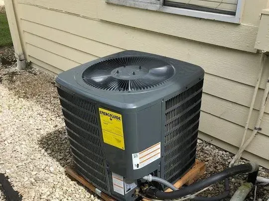 Outdoor air conditioner unit beside a beige house wall, with black hoses and gravel ground.