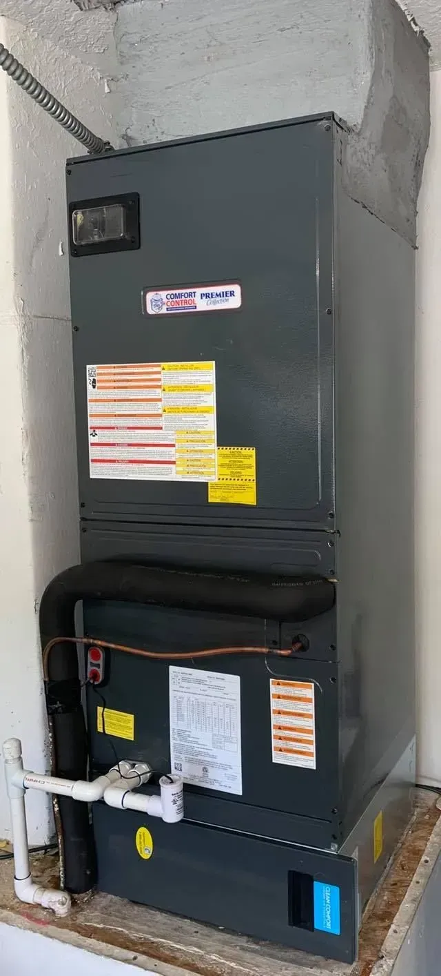 Gray HVAC furnace and AC unit stacked in a utility closet with labels and exposed pipes.