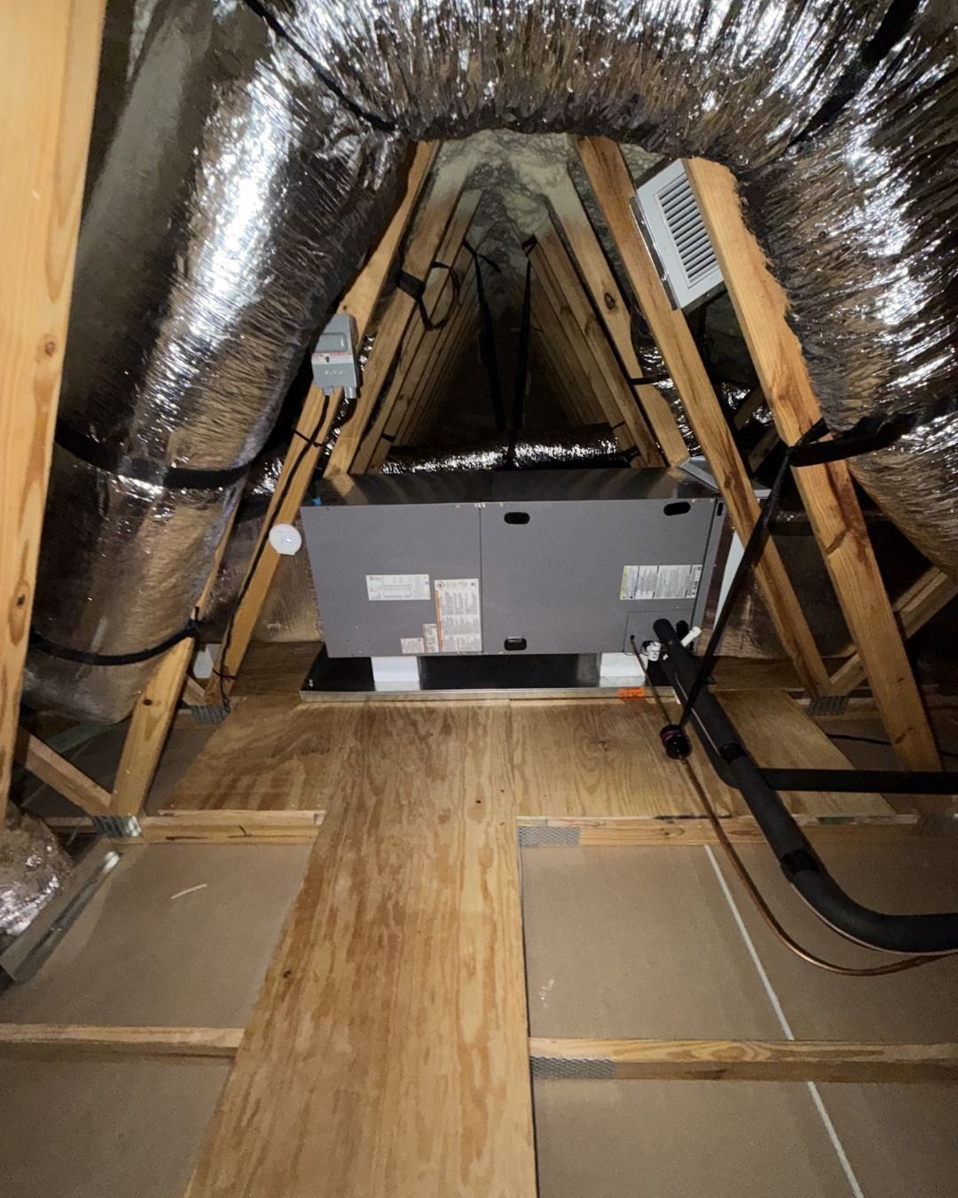 Attic interior with exposed ductwork and a large HVAC unit on the floor, seen through wooden rafters.