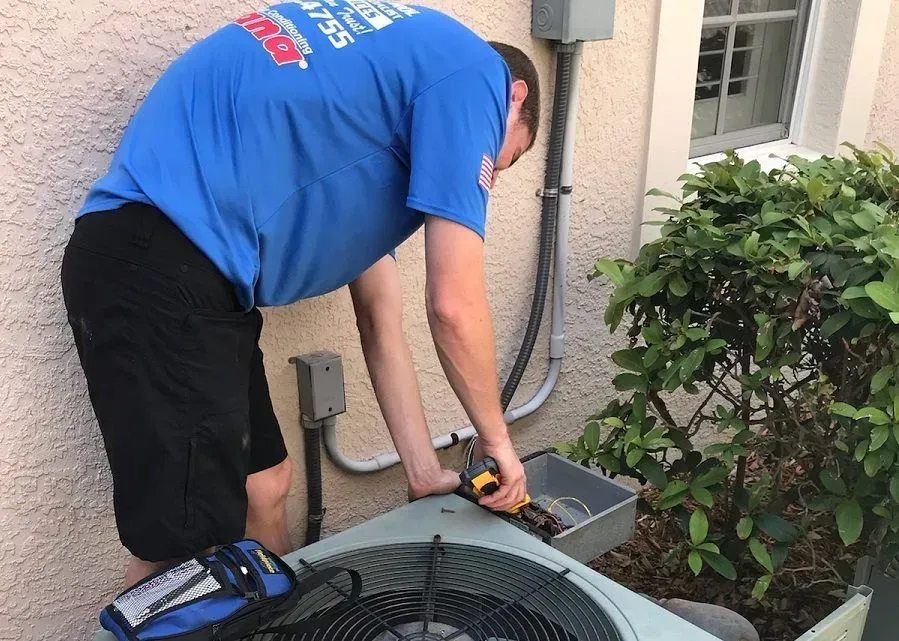 Technician in blue shirt servicing an outdoor HVAC unit beside a house