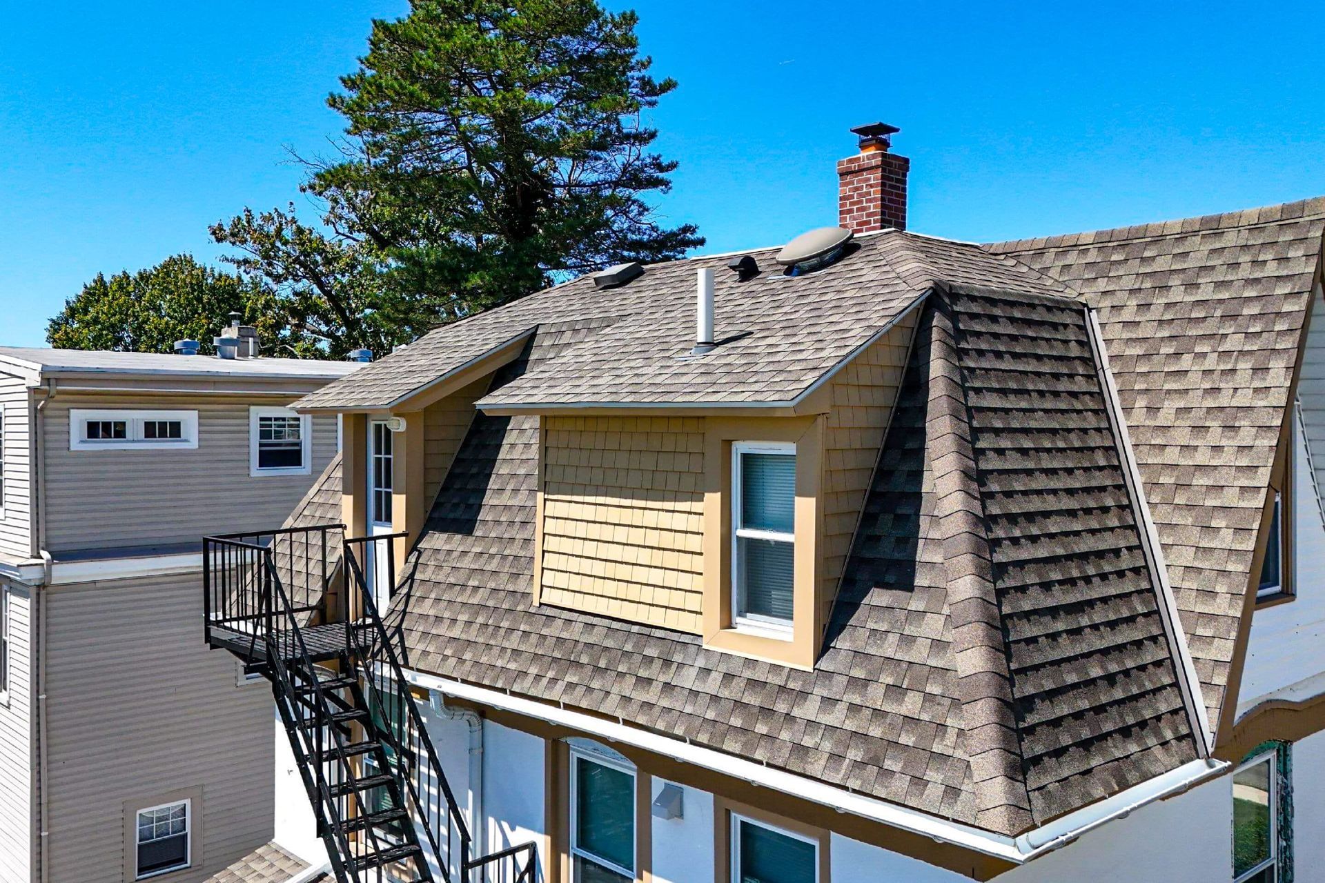 An exterior view of a multi-story house featuring a brown-shingled roof, a dormer, and a black metal fire escape.