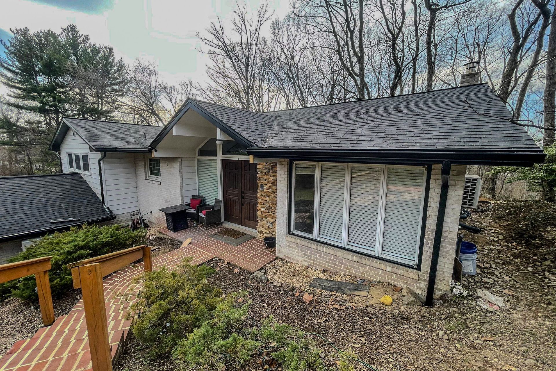 A single-story house with a white textured exterior, a dark shingled roof, a brick walkway, and large front windows.