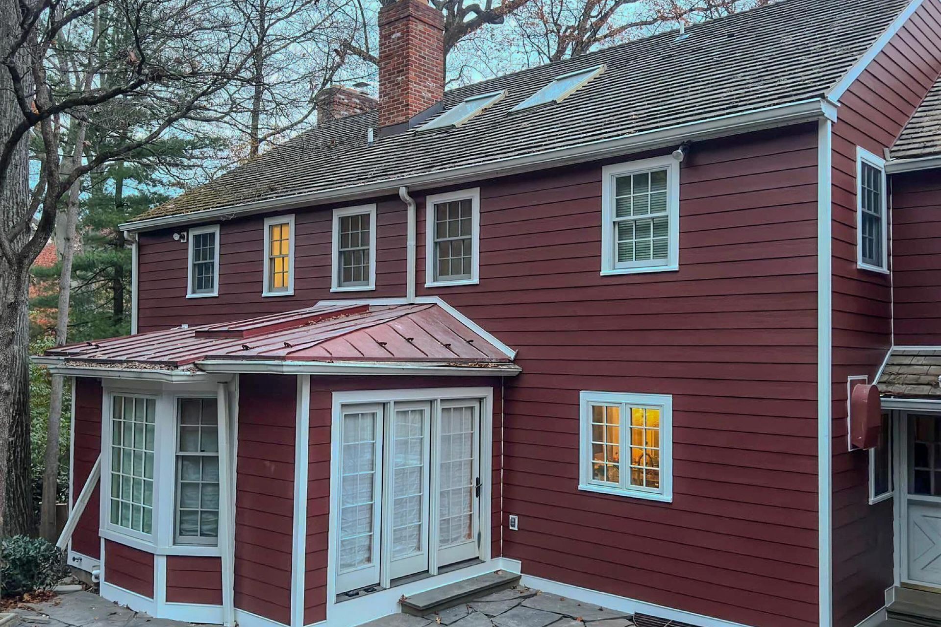 A two-story, maroon-colored house with white trim, a metal roof, and several windows nestled among trees.