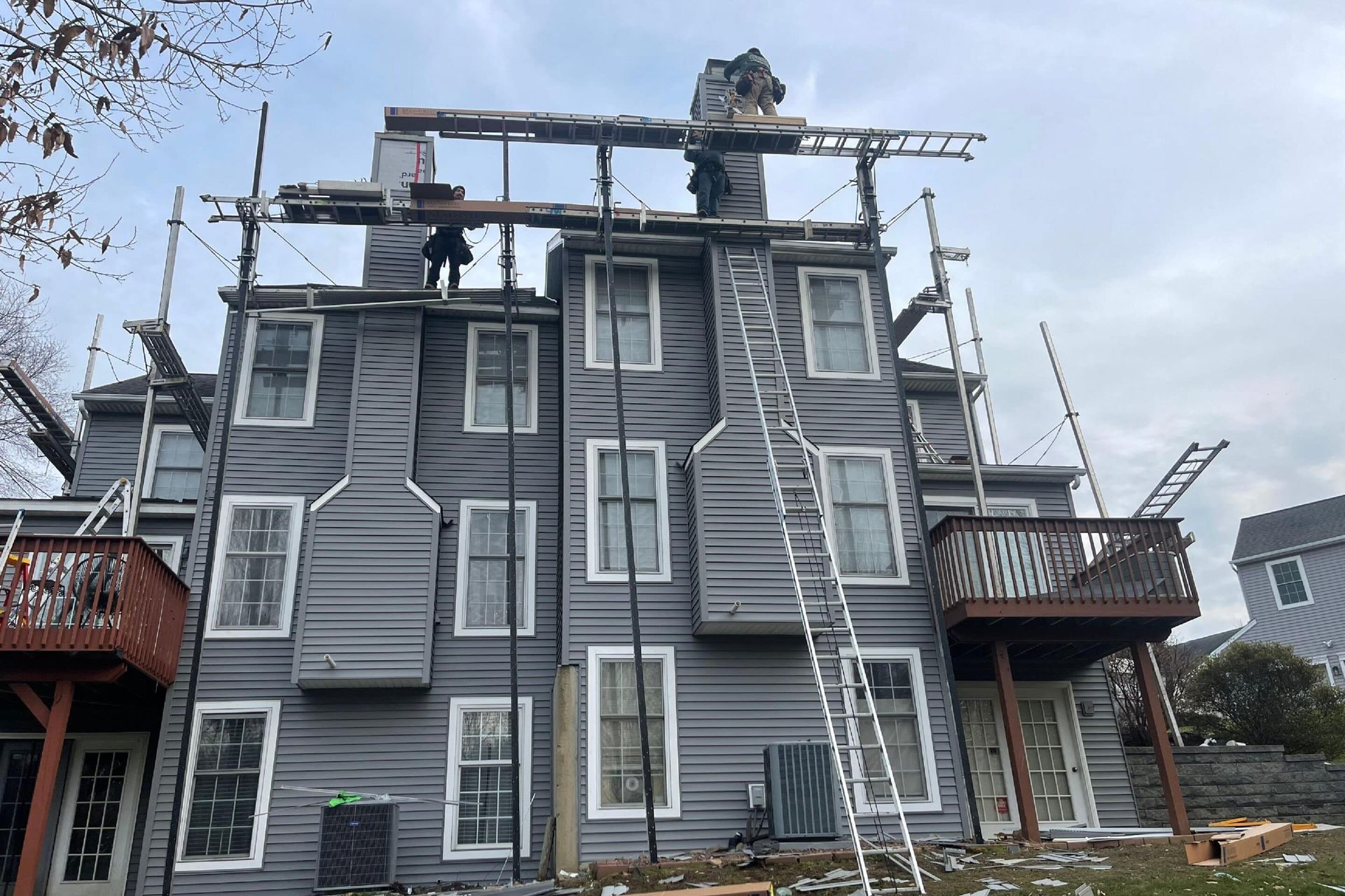 Gray residential building under repair with external scaffolding and a worker atop a ladder near the chimney.