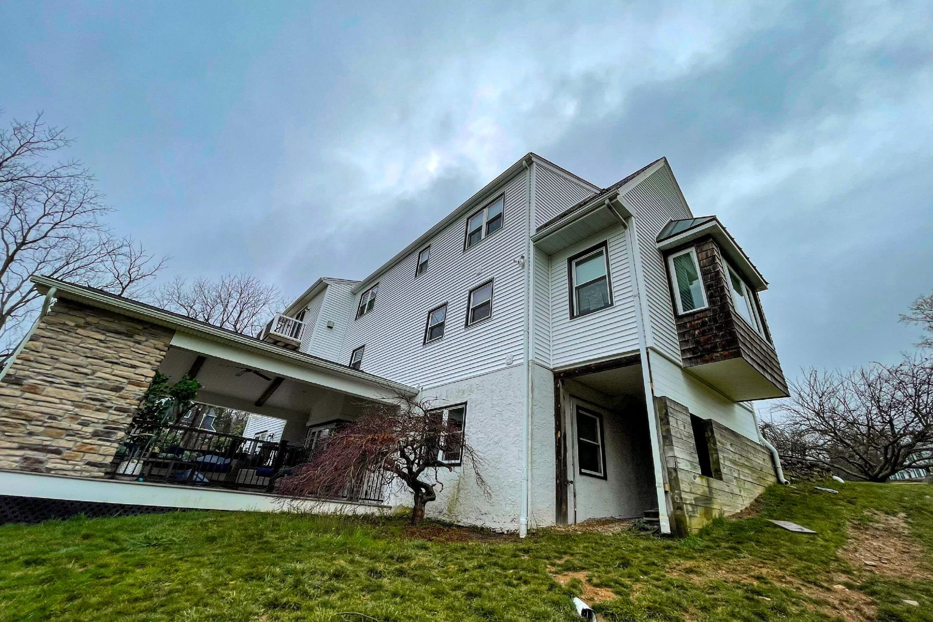 A low-angle view of a multi-story house featuring white siding, a stone facade, a covered patio, and a bay window.
