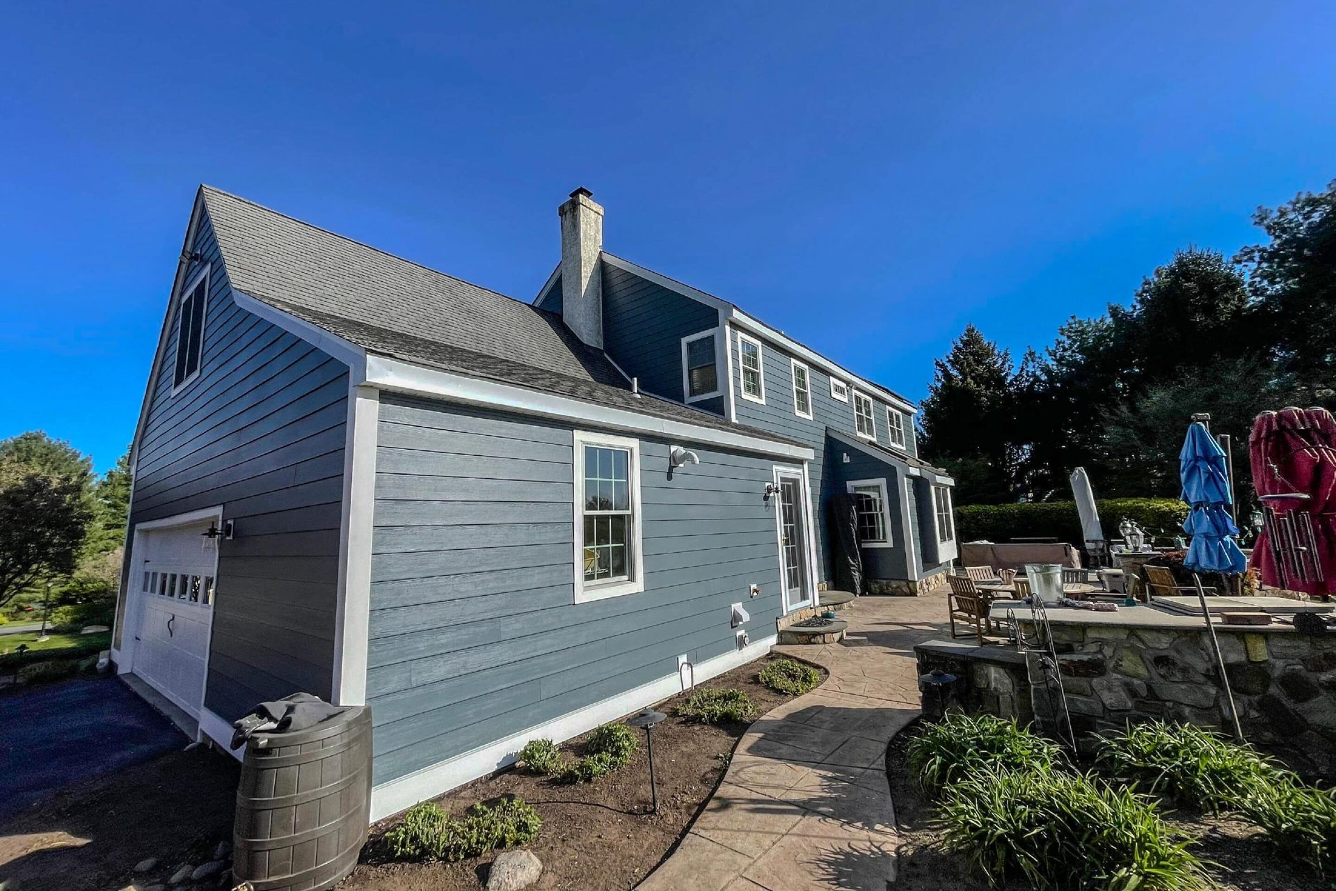A blue, two-story house with a white-trimmed garage and chimney sits under a clear, bright blue sky next to a patio.