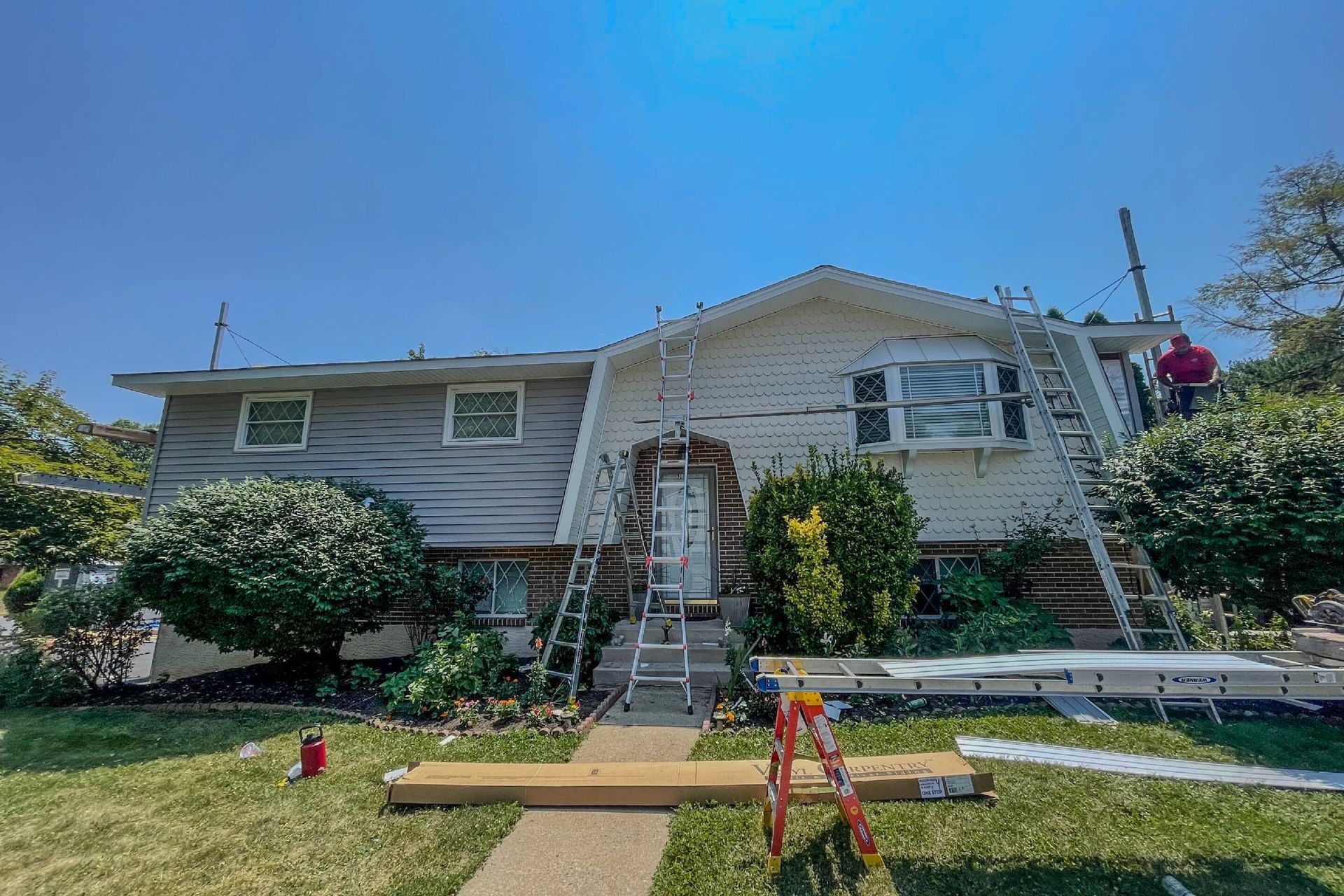 A two-story house under renovation with scaffolding, ladders, and siding materials in the front yard under a blue sky.