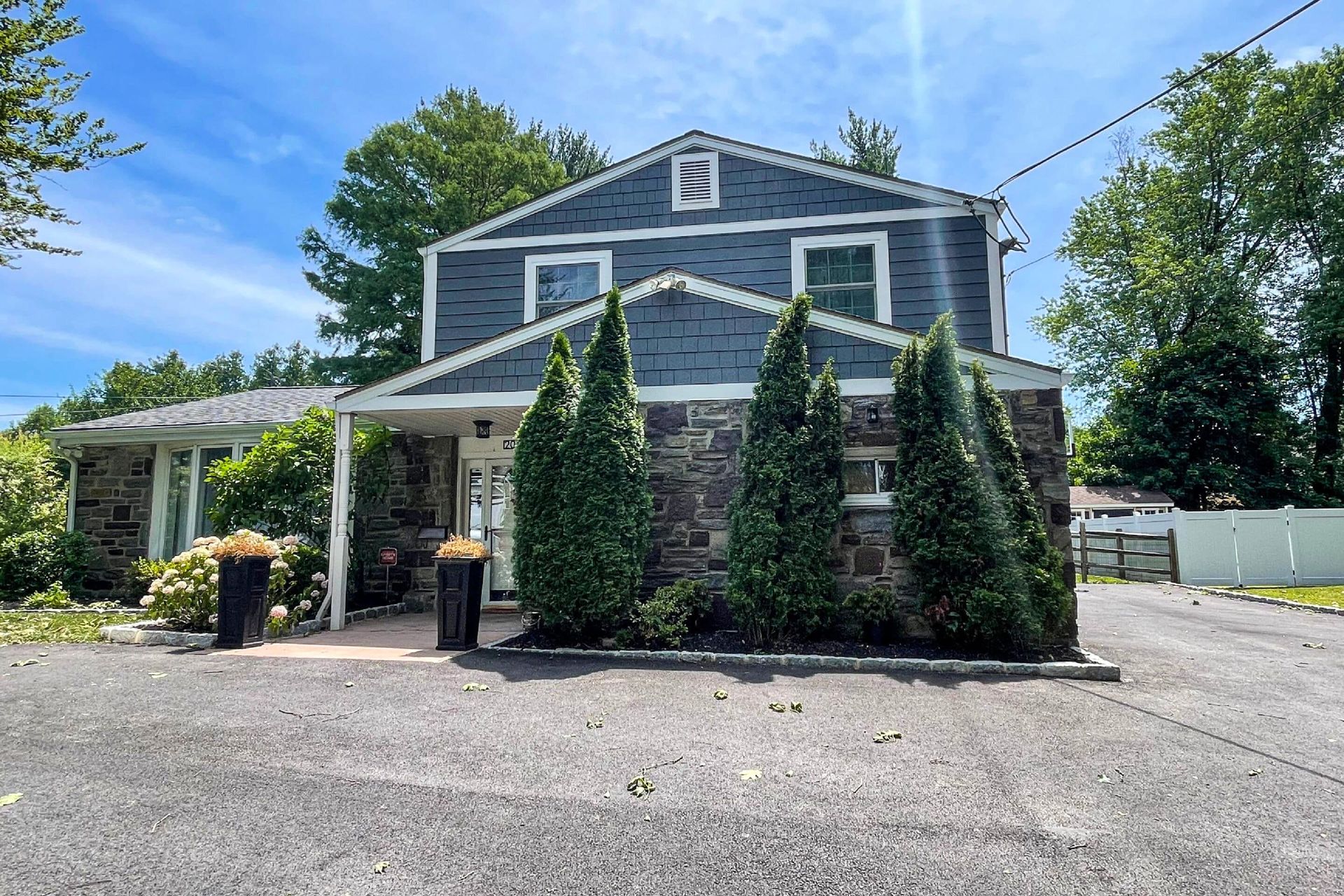 A two-story house with dark grey siding and stone masonry, featuring three tall evergreen trees in the front yard.