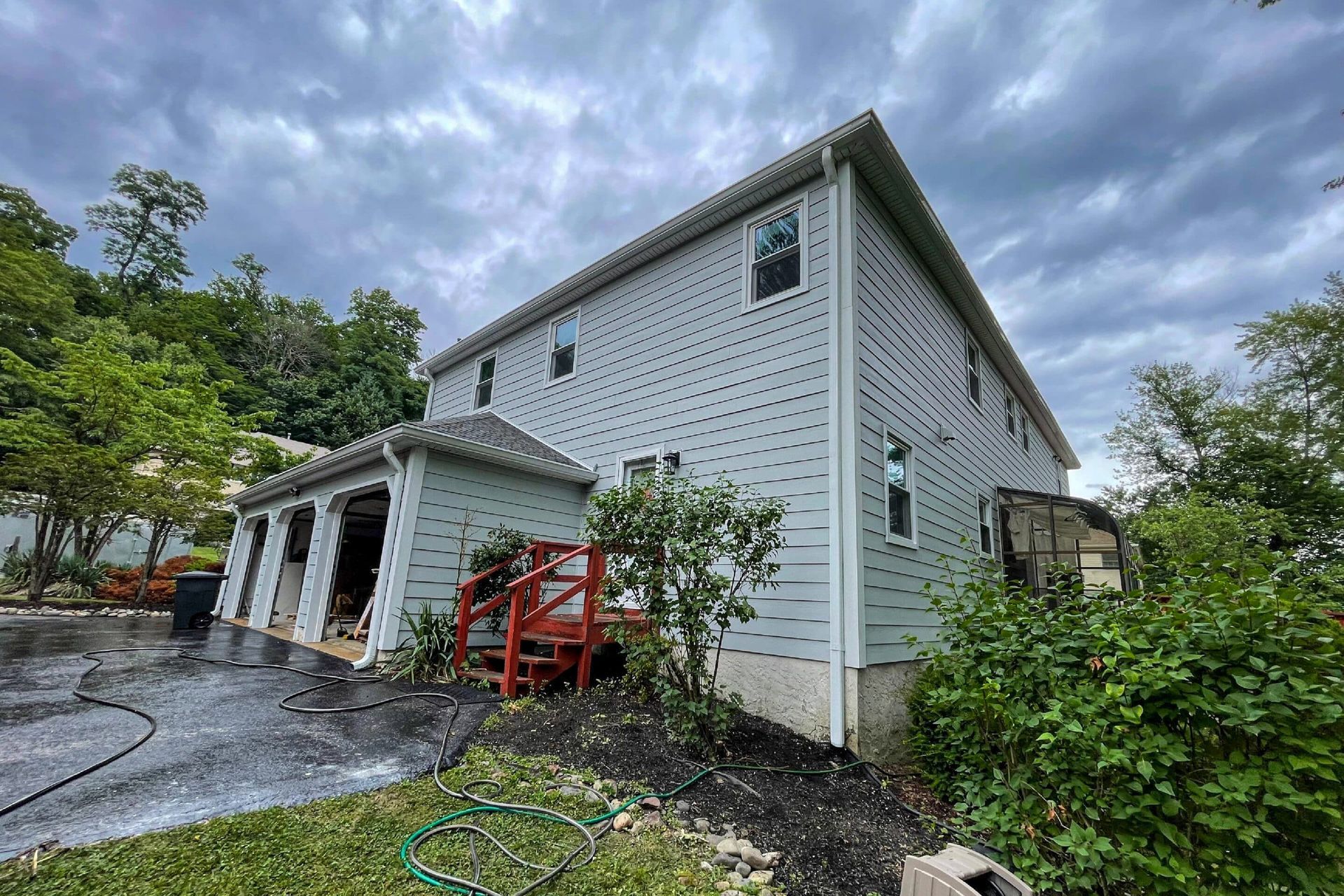 A light-gray, two-story house with a white-trimmed garage and red wooden stairs under a cloudy, overcast sky.