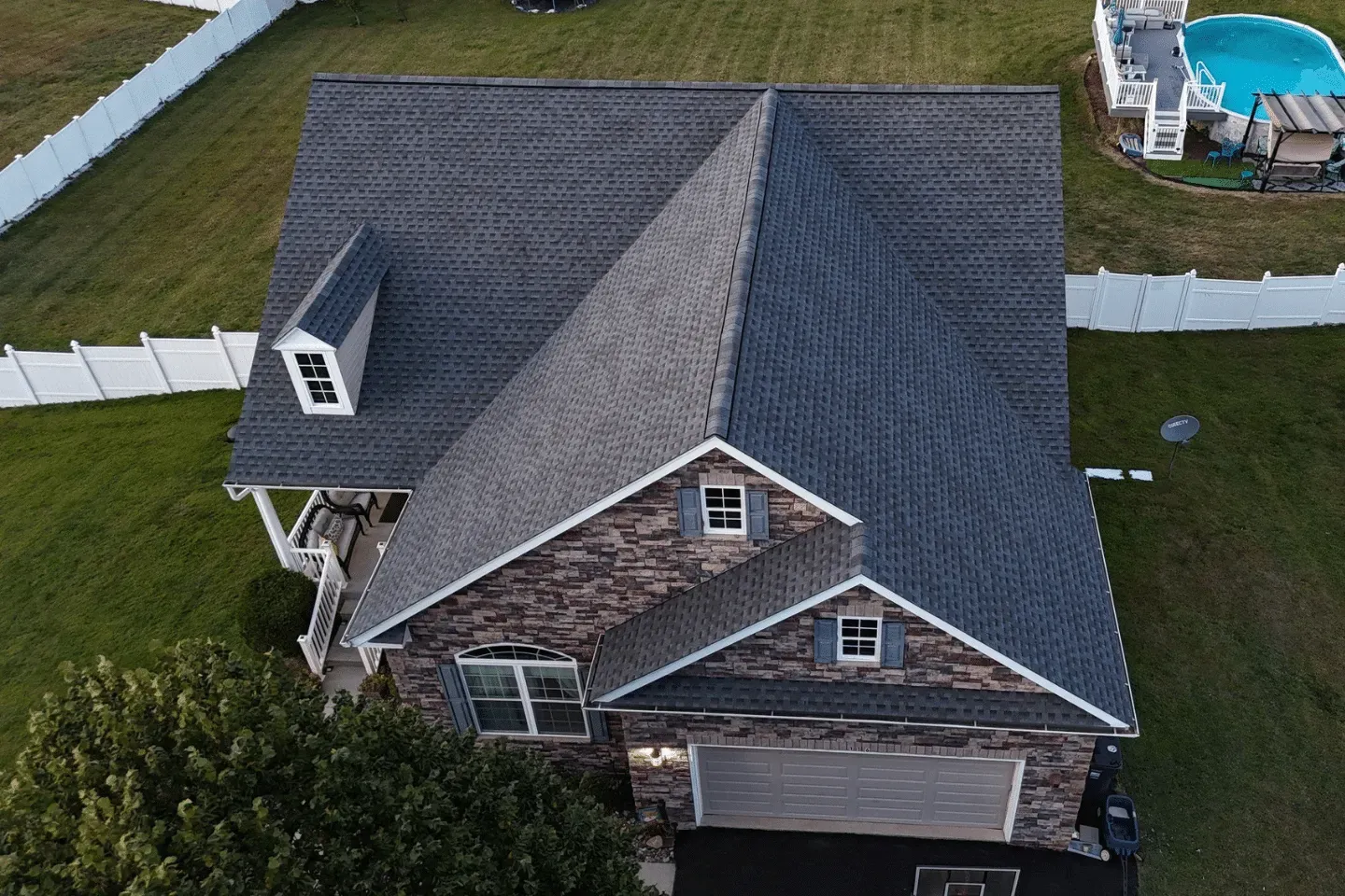 Aerial view of a gray shingled roof on a stone-faced suburban house with a yard, white fencing, and a swimming pool.