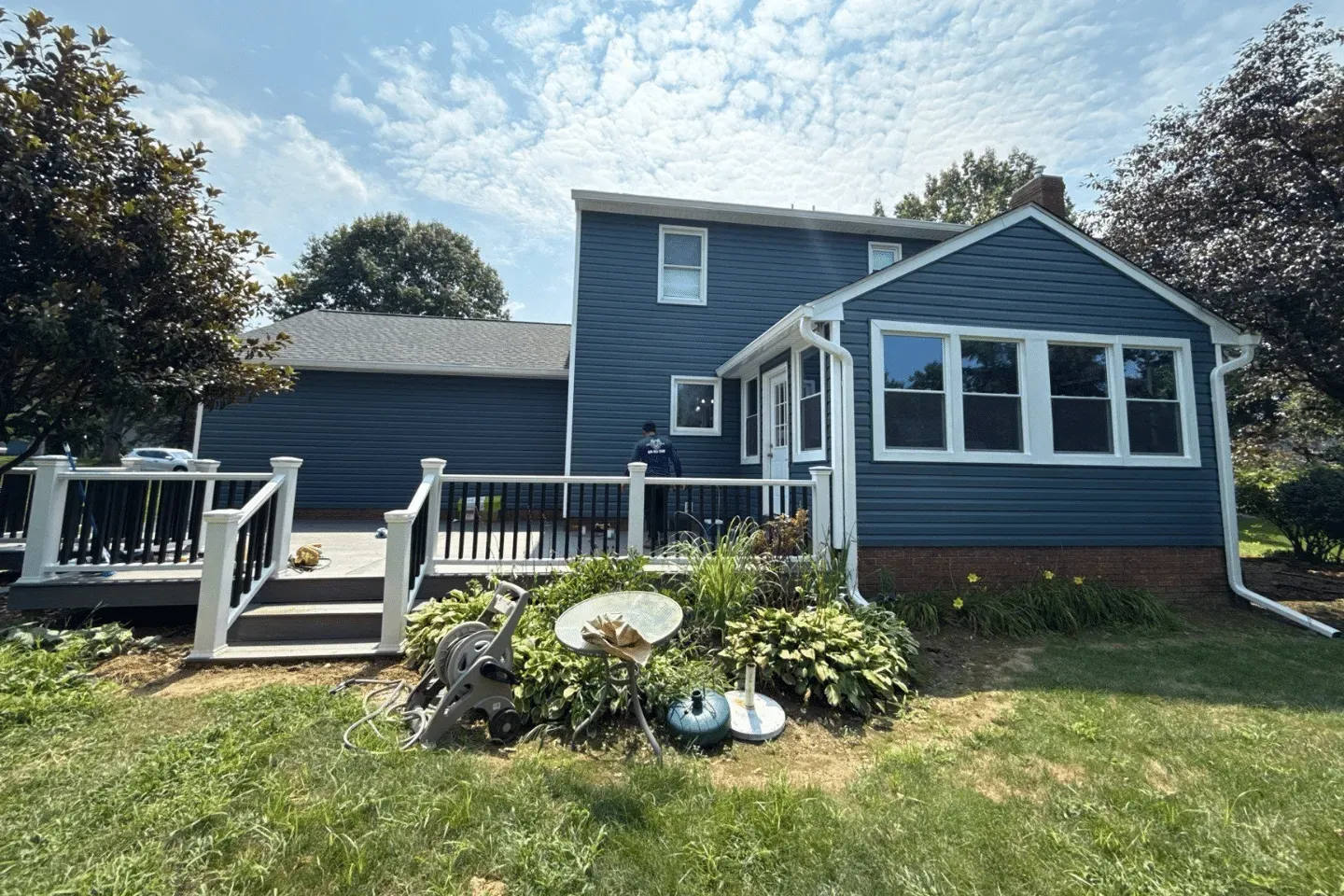 A blue two-story house with a wooden deck, a sunroom with white windows, and a landscaped yard.