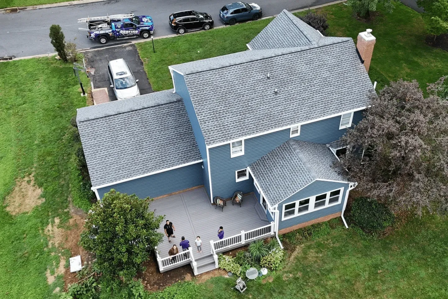 High-angle view of a blue, two-story house with a grey deck, driveway, and a small group of people standing on the deck.