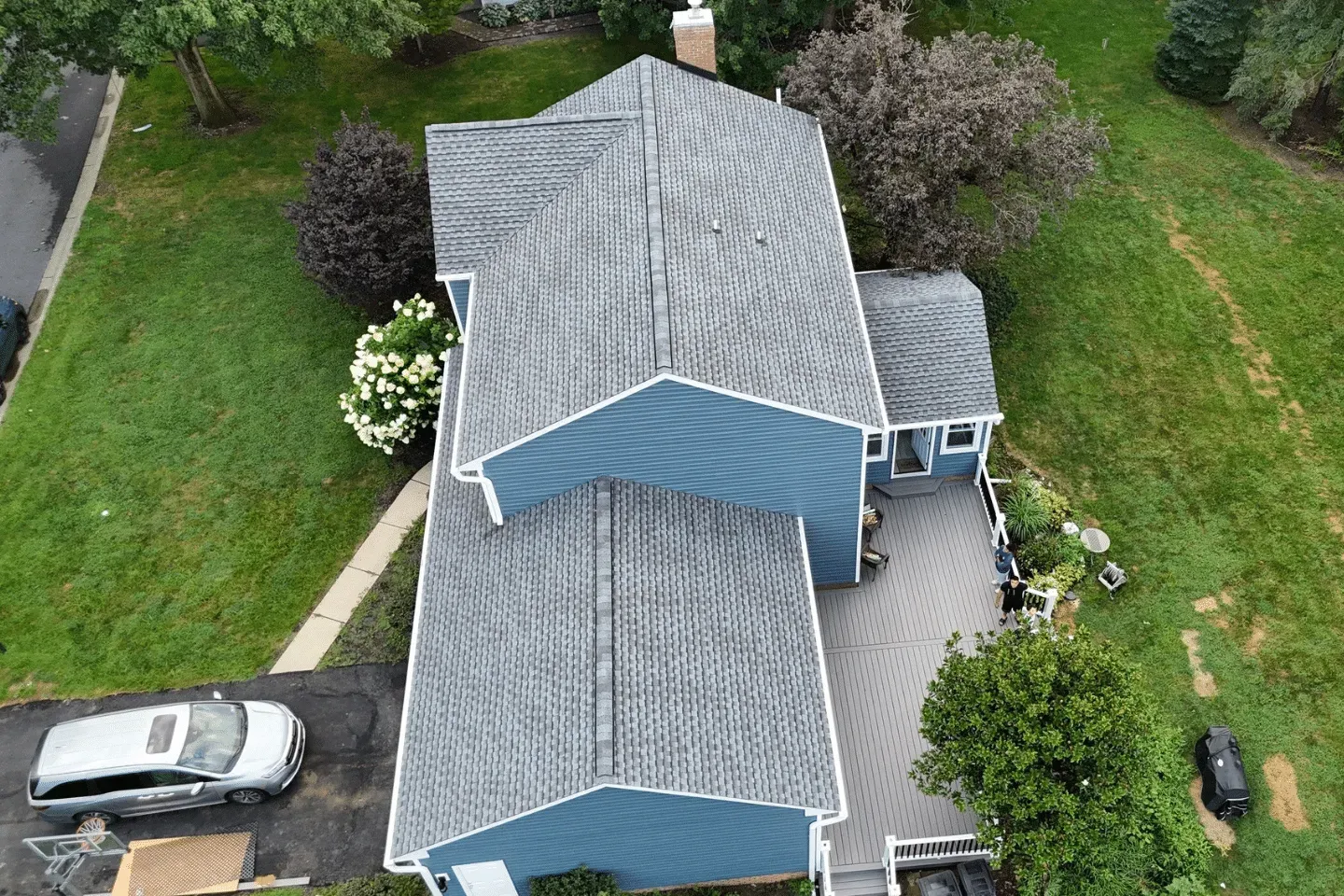 A high-angle aerial view of a blue house with a gray shingled roof, a gray patio, and a driveway with a white car.