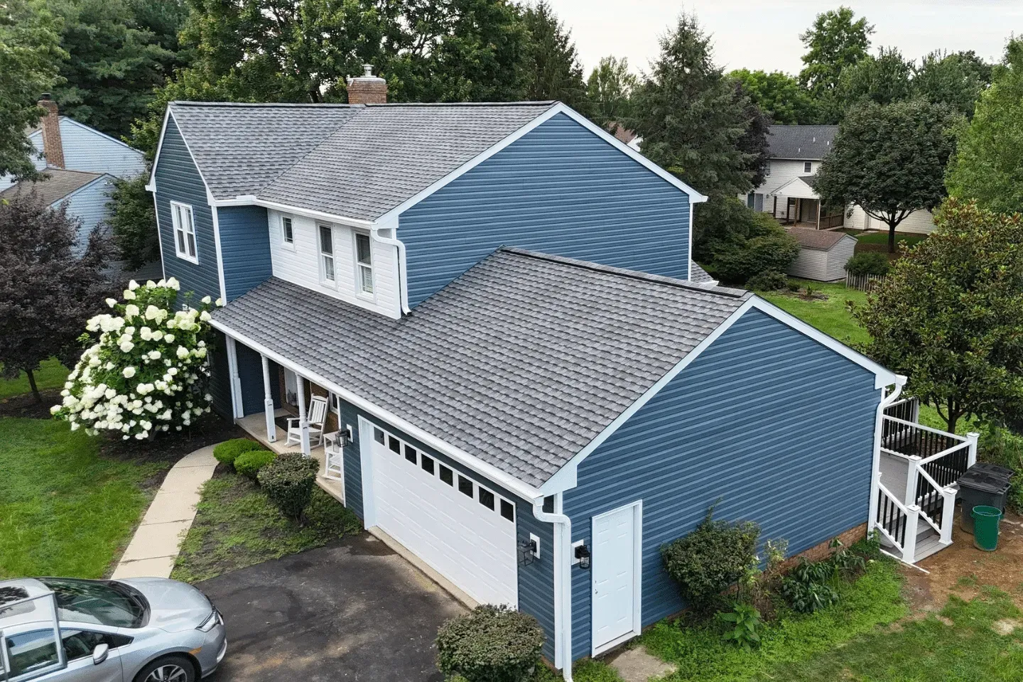 Aerial view of a two-story blue-sided suburban house with a gray roof and attached garage, surrounded by green trees.