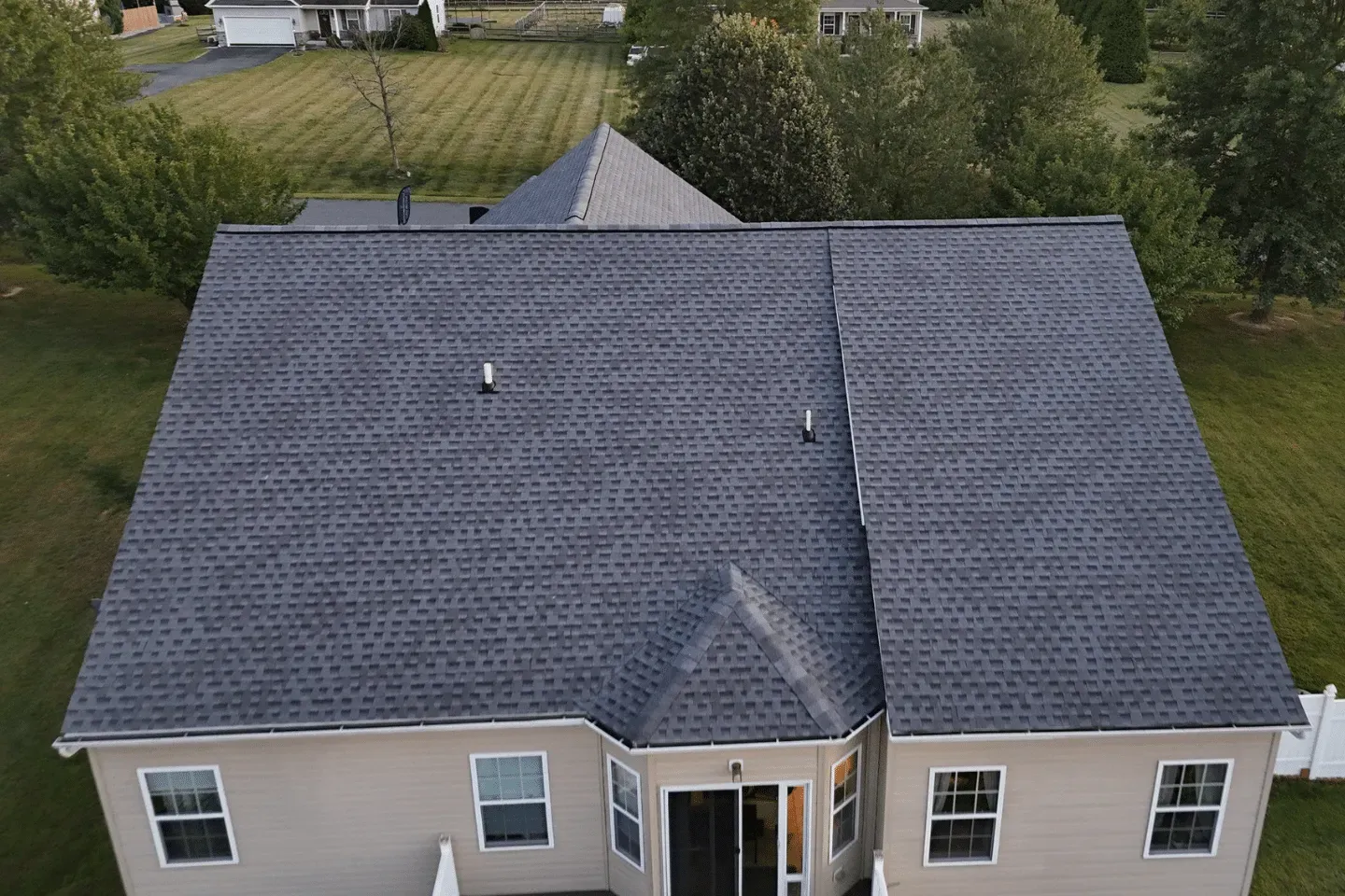 Aerial view of a beige house with a dark gray shingled roof, set in a green backyard.
