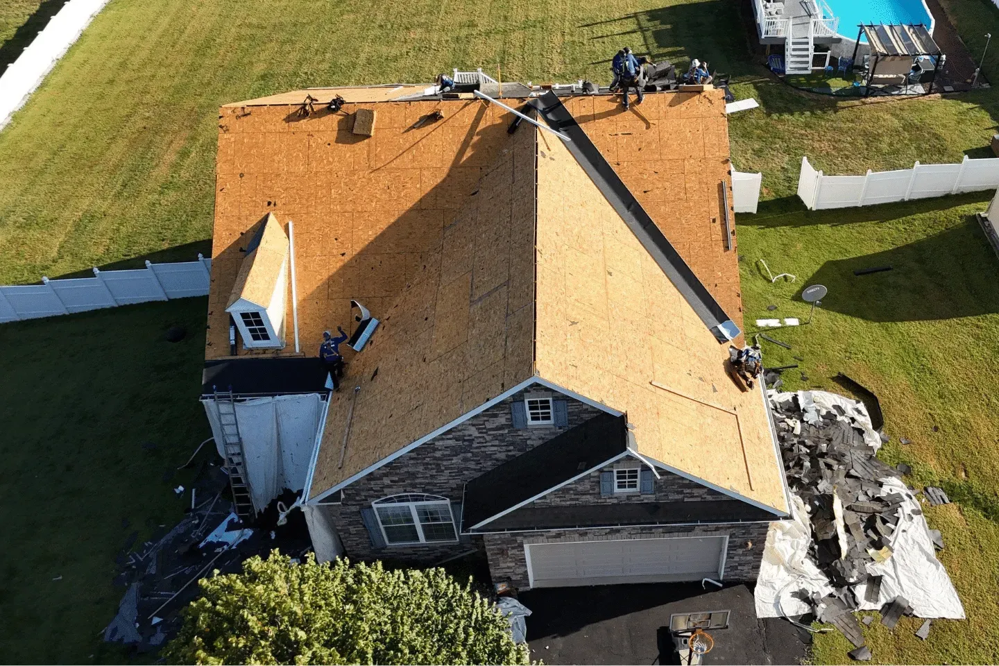 Aerial view of workers replacing shingles on a house roof with exposed plywood sheathing and debris on the ground.