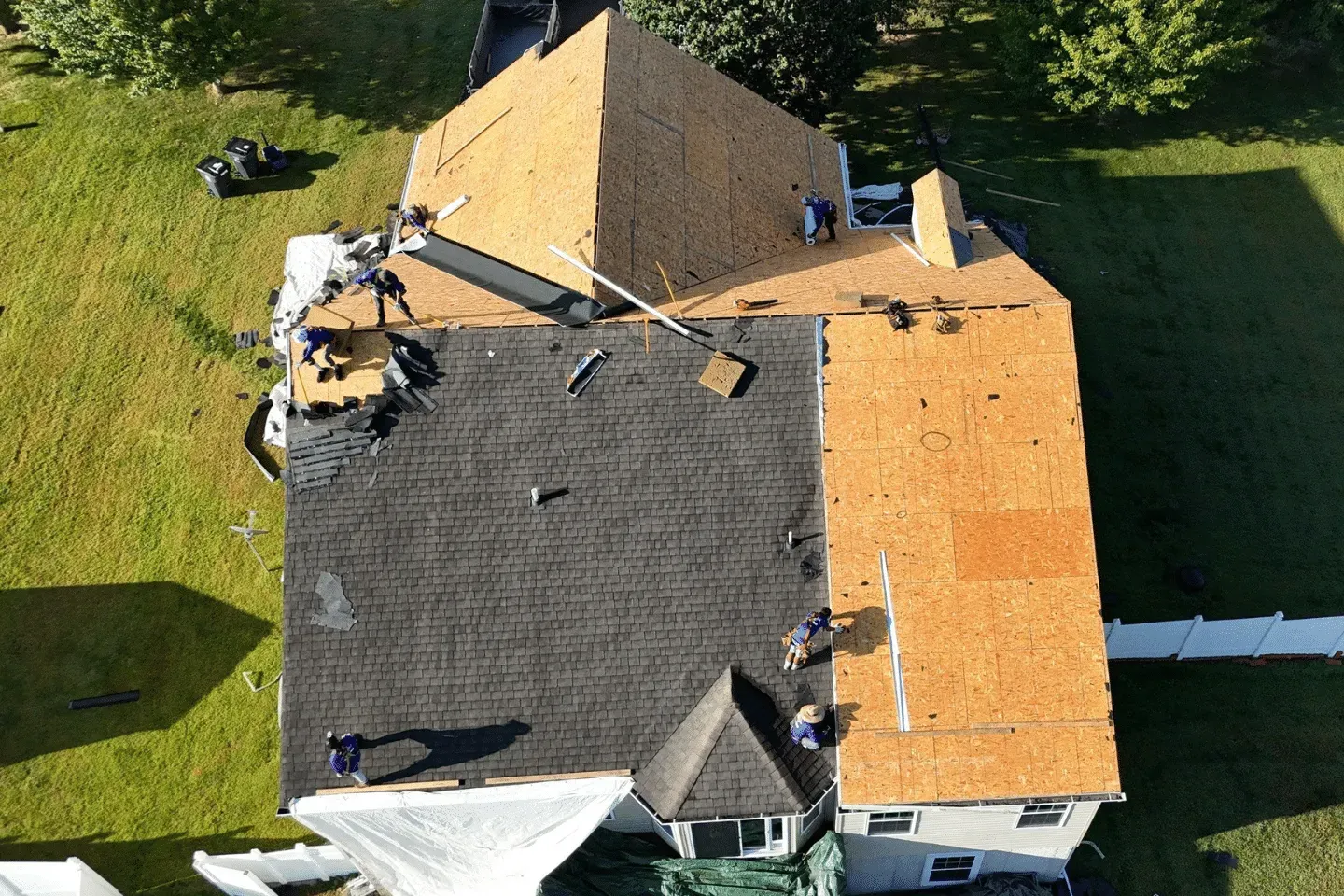Aerial view of workers replacing a residential roof, showing sections of exposed plywood and new black shingles.
