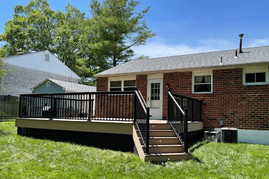 A brown brick house with a newly built tan deck, black railings, and stairs leading to the backyard under a clear sky.