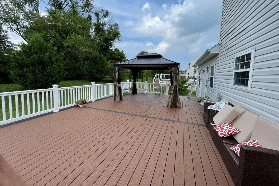 A spacious brown composite deck features a dark gazebo and a long outdoor sofa with patterned pillows against a house.