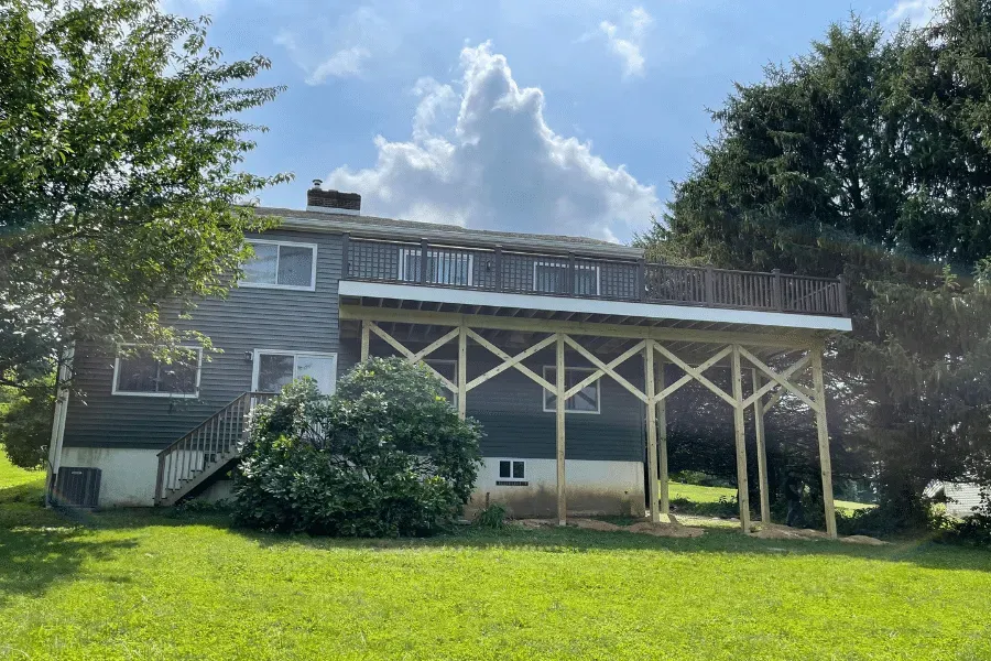 A two-story house with dark siding and a large wooden deck supported by multiple diagonal support posts over a lawn.