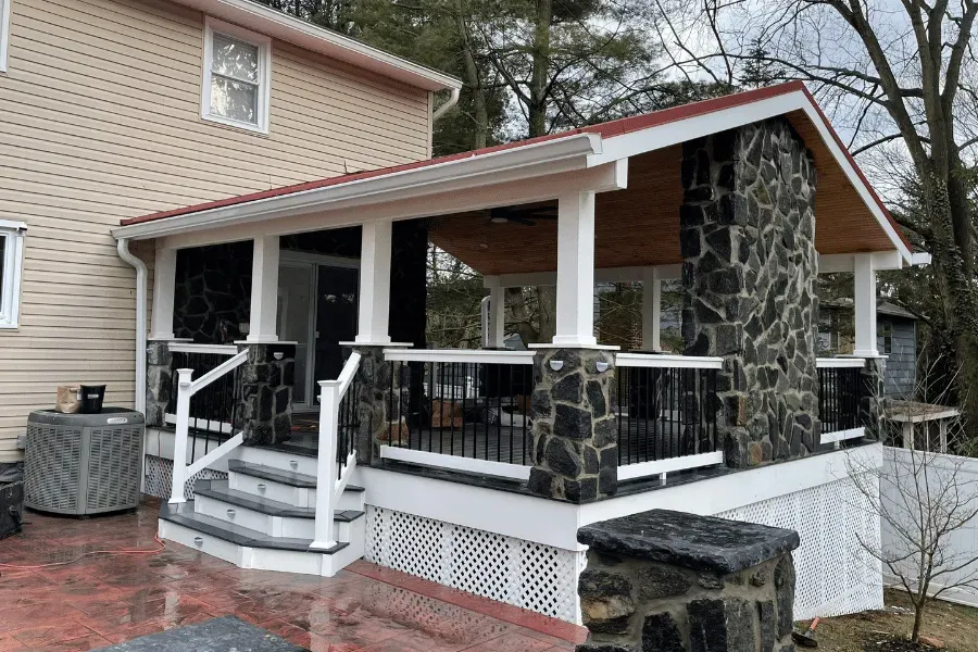 A raised wooden deck with white railings, stone pillars, a stone fireplace, and a covered porch attached to a house.