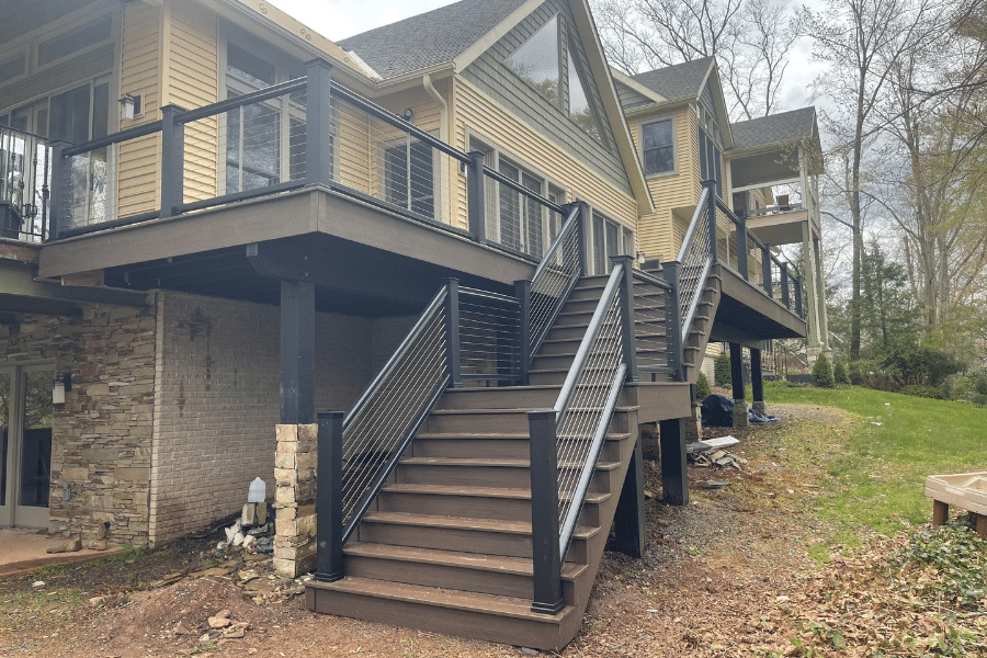 A tan house with a raised wooden deck featuring black railings and stairs leading down to a wooded backyard.