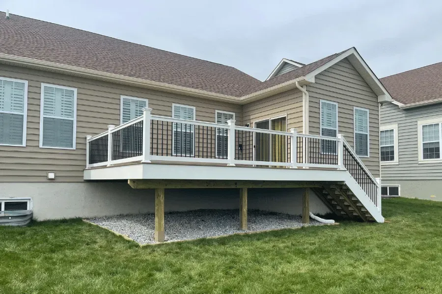 A raised wooden deck with white railings and stairs attached to the back of a beige siding house with a gravel base.