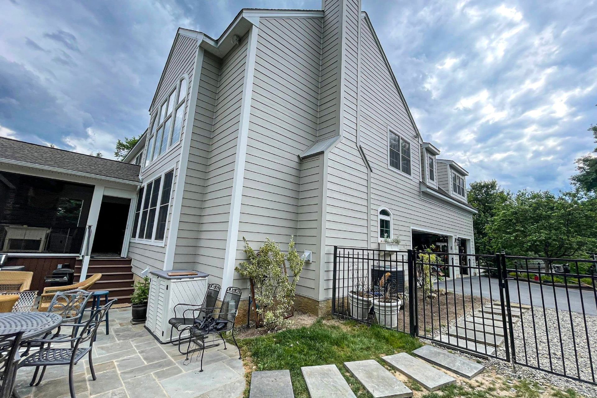 Exterior view of a light gray, multi-story home with a patio, metal fencing, and a garden against a cloudy blue sky.