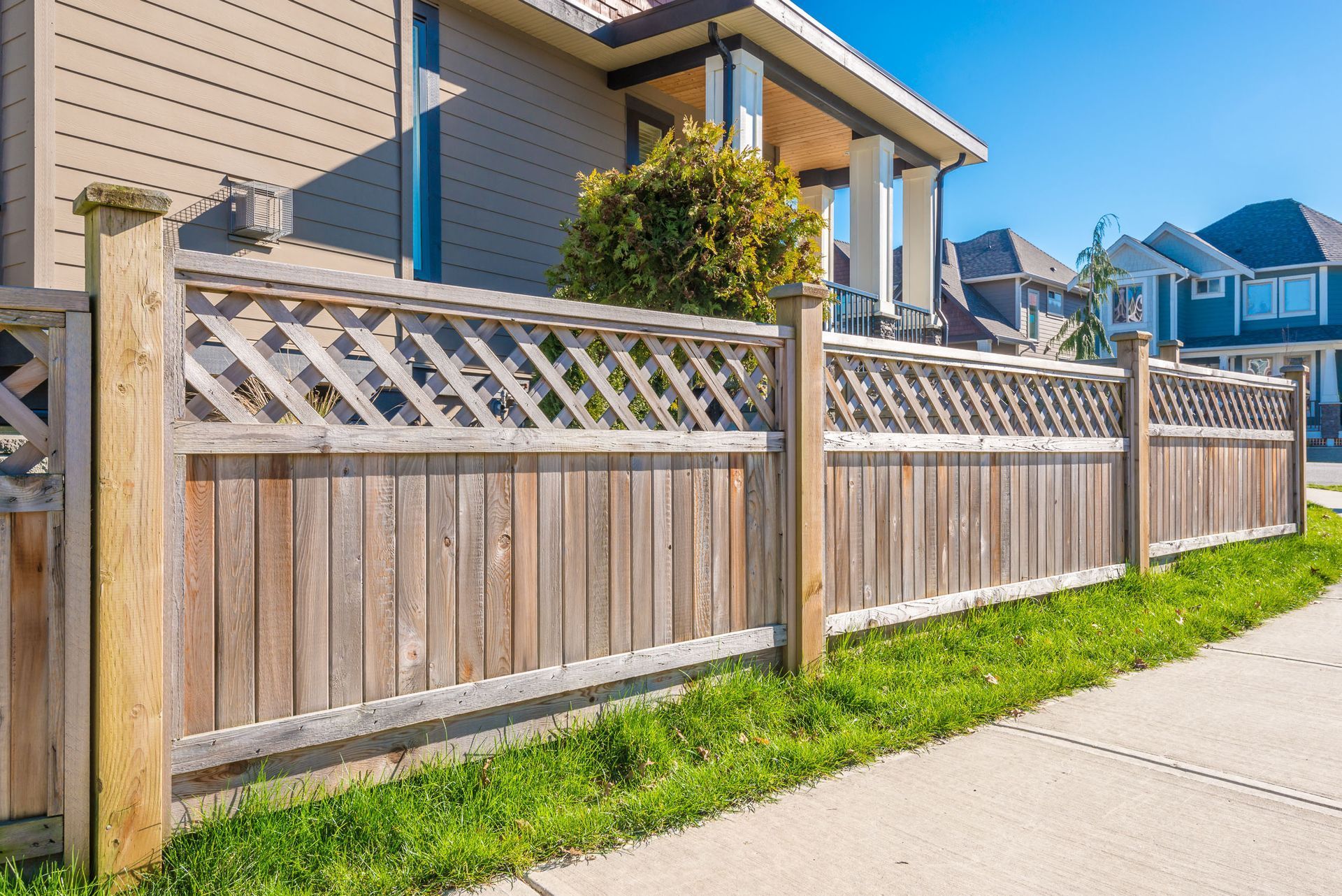 Wooden fence with lattice top, separating a lawn from a sidewalk, houses in the background.