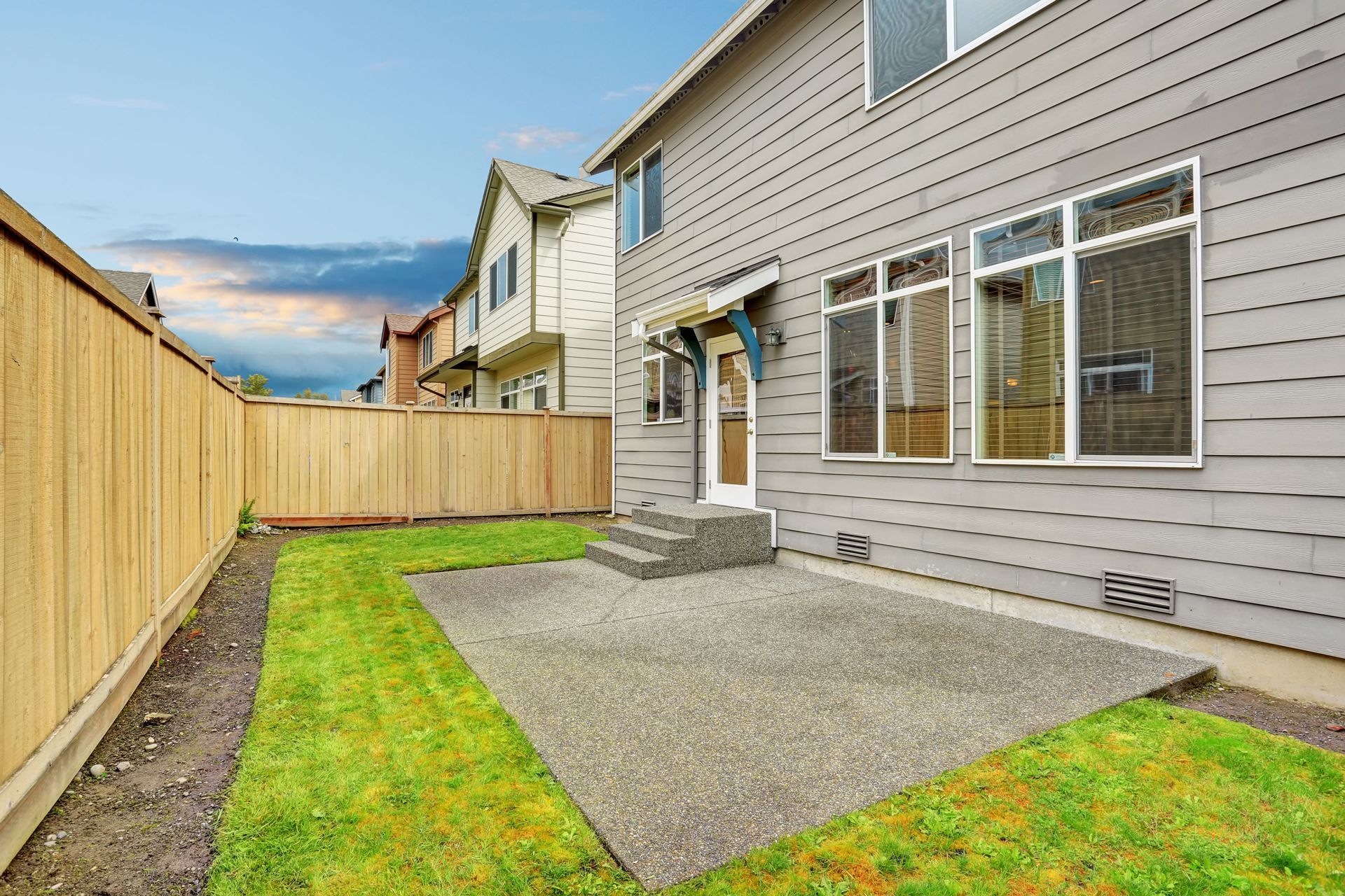 Backyard with concrete patio, grass, wooden fence, and gray house with windows.