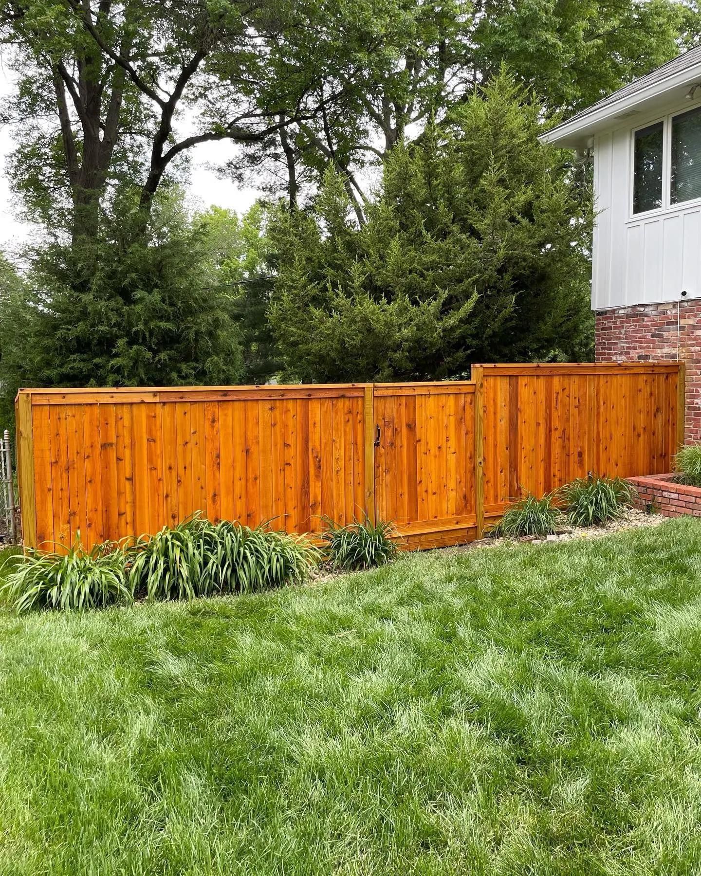 Wooden fence with reddish-brown stain on a green grassy lawn near a white house with a brick foundation.