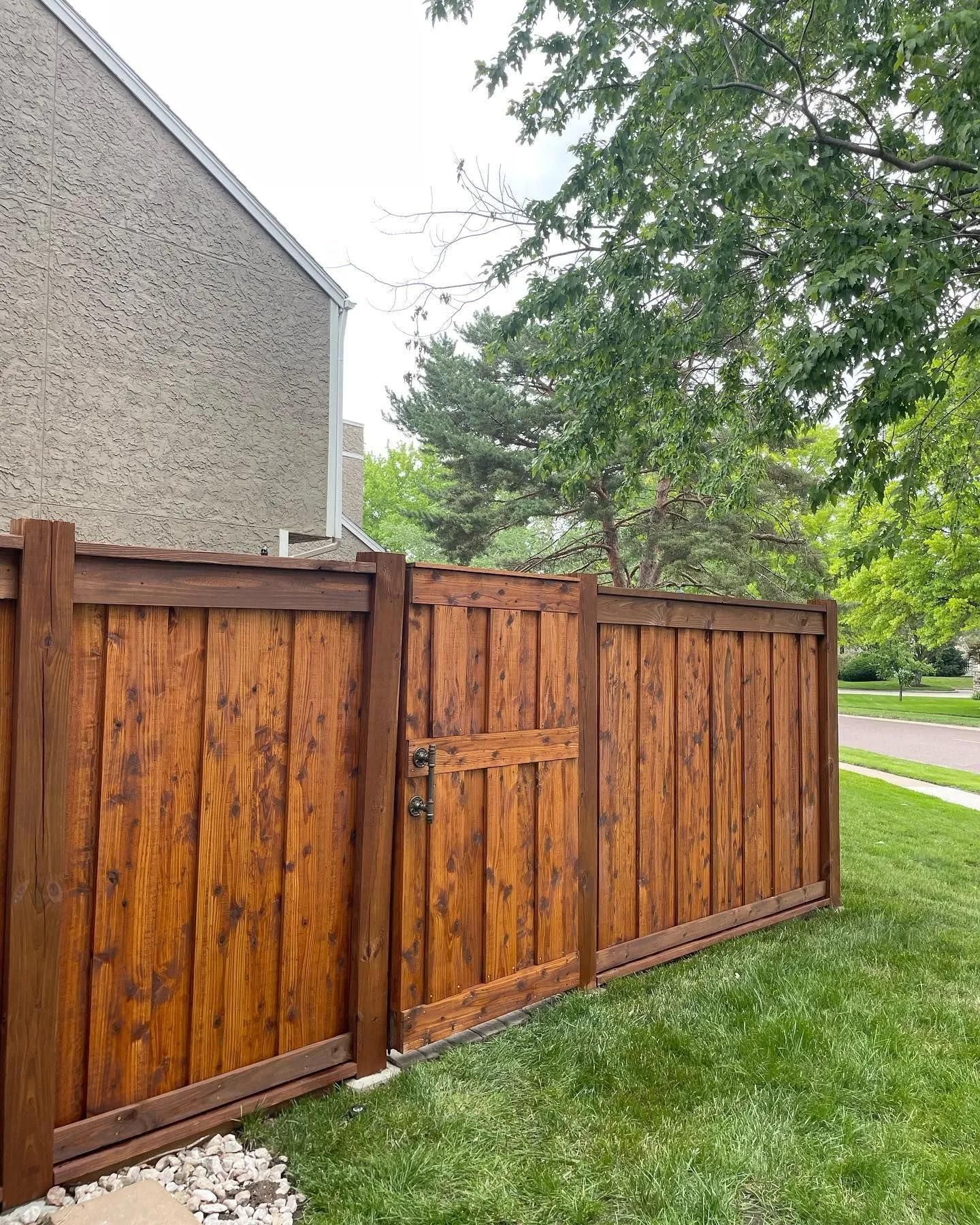Wooden fence with gate; stained brown, bordering a grassy lawn next to a stucco building and a green tree.