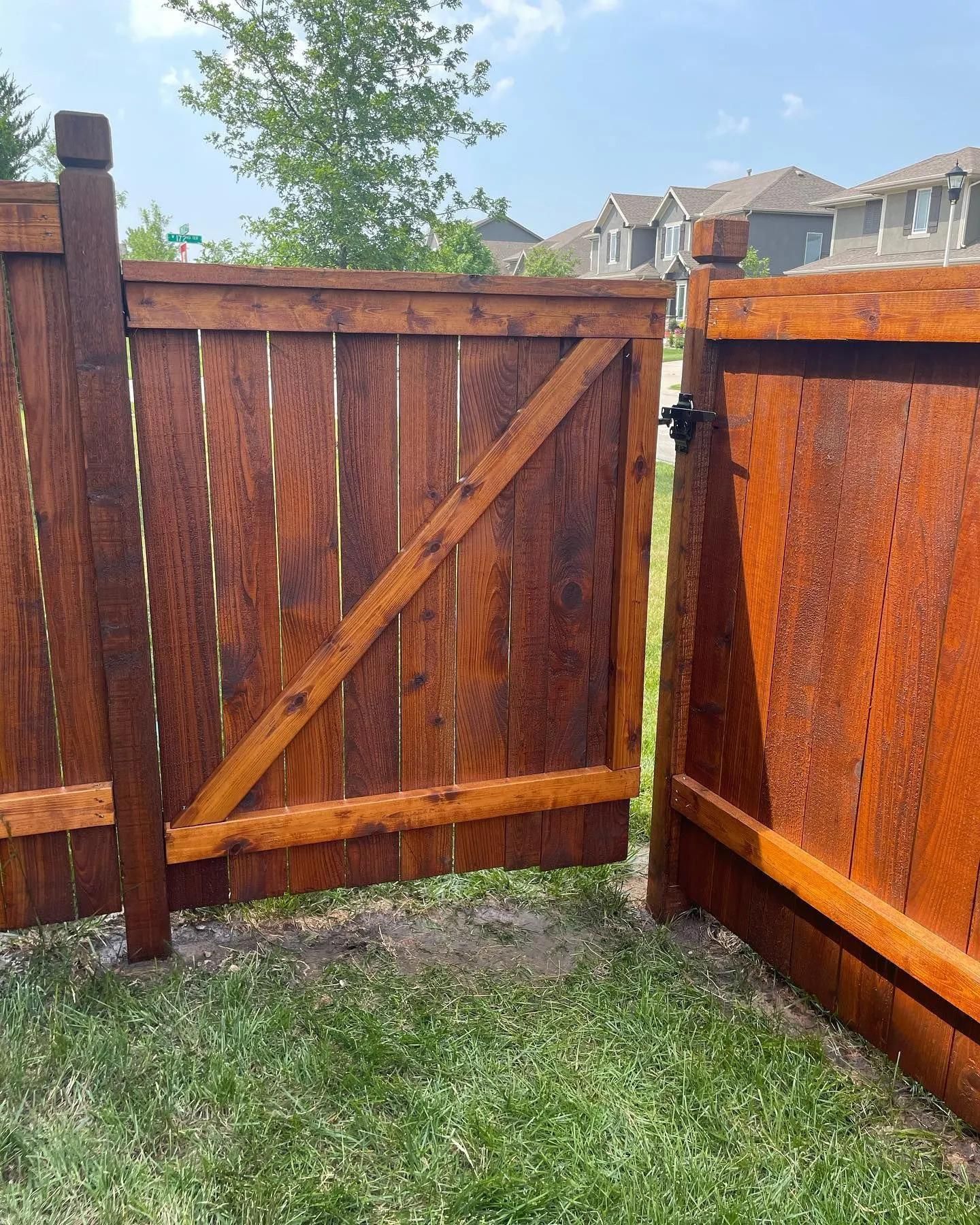Wooden gate in backyard, stained brown, with diagonal support. Grass and houses in the background.