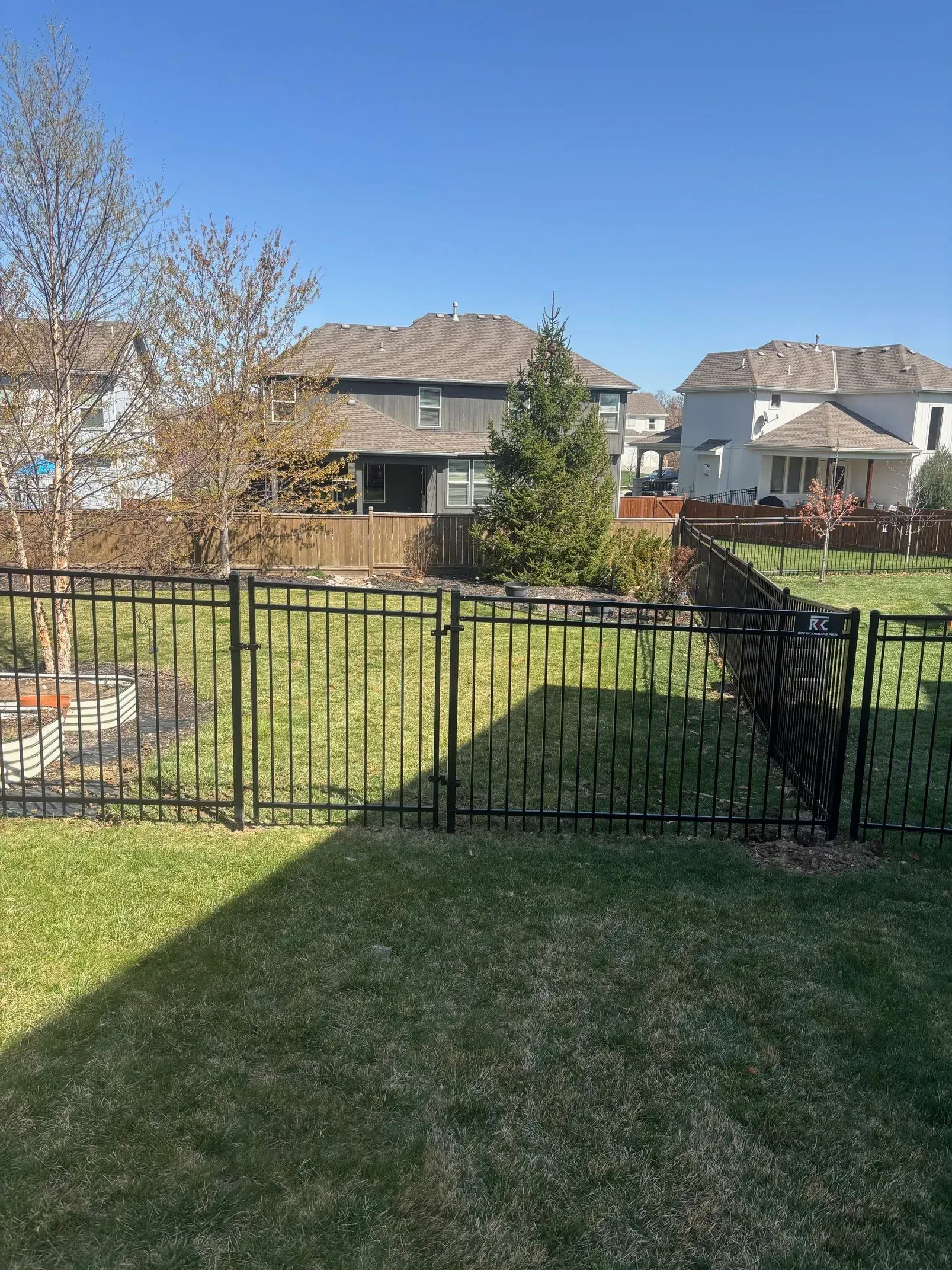 Black fence in backyard with green grass, houses, and trees under a blue sky.