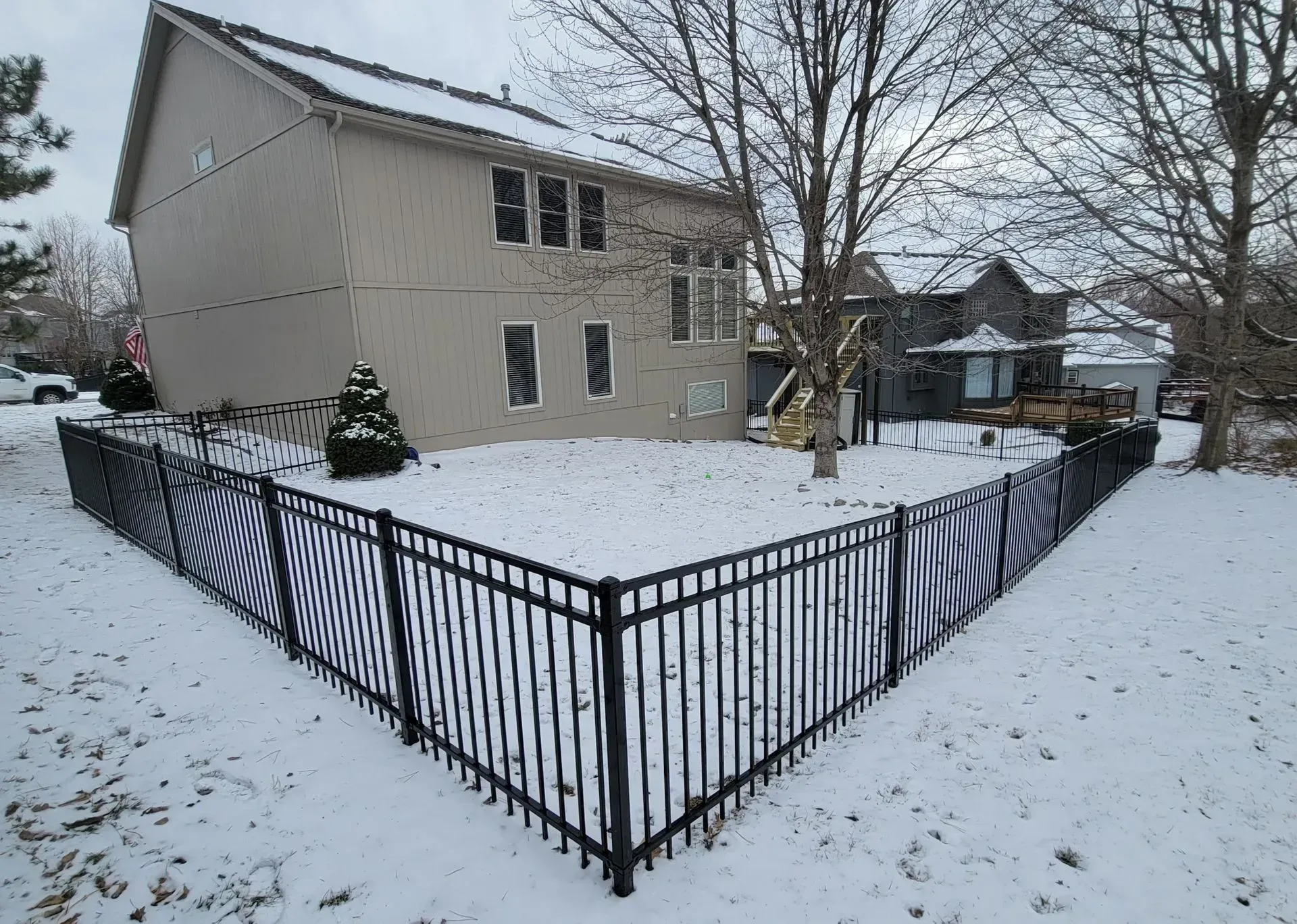 Black metal fence surrounds a snow-covered backyard of a two-story beige house. Winter setting with trees.