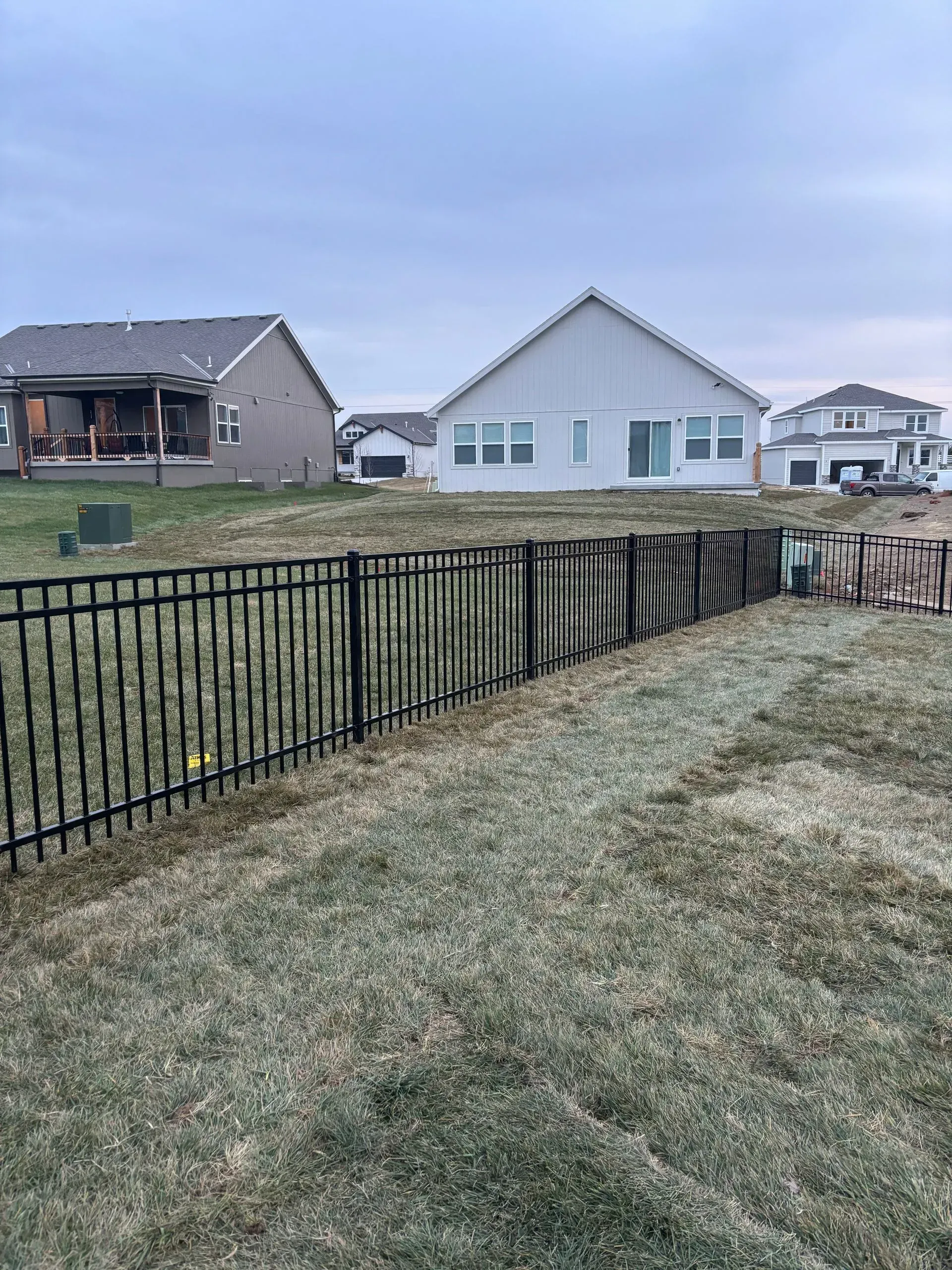 Black metal fence along a grassy slope, with houses in the background under a cloudy sky.