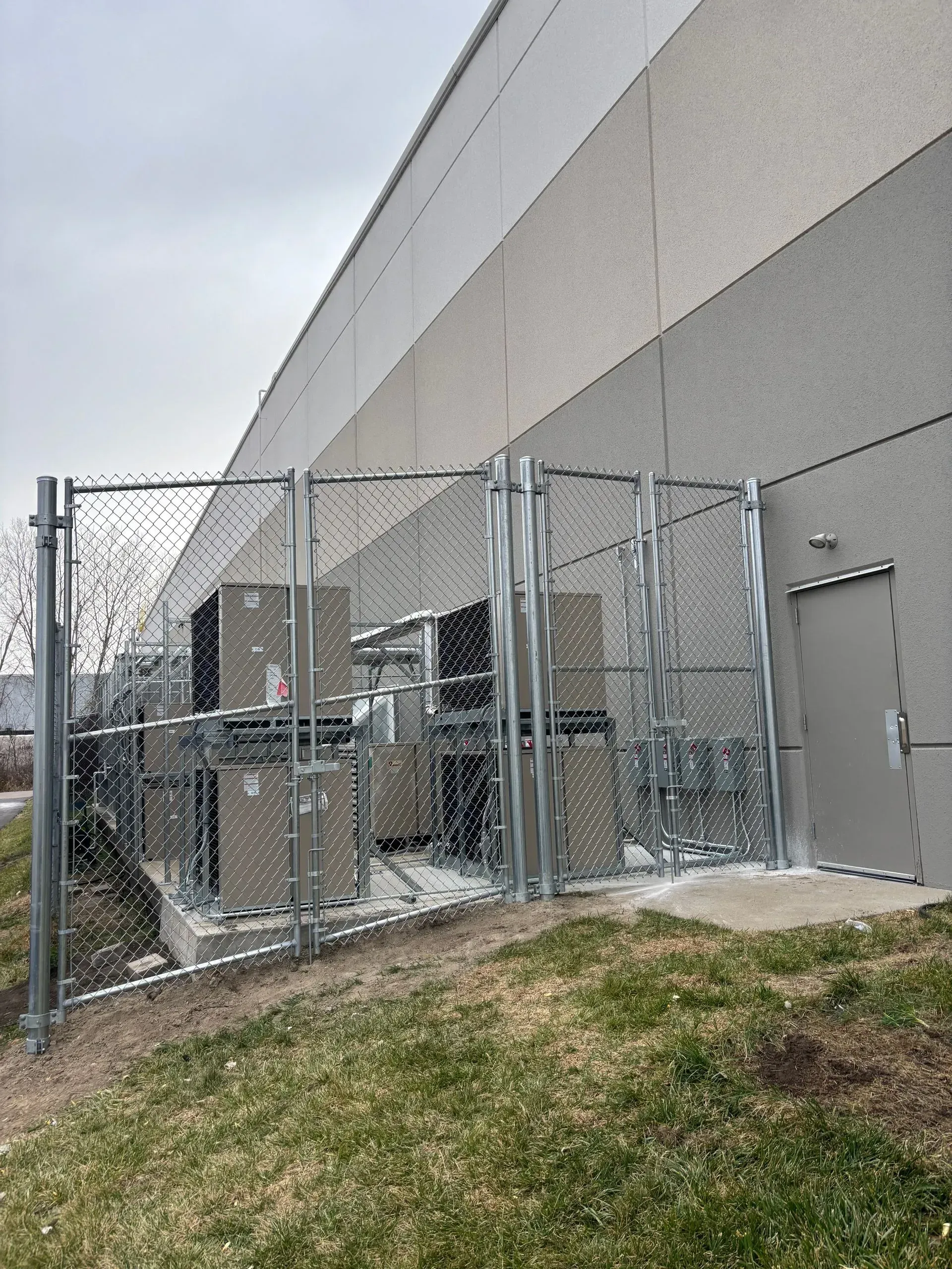 Chain-link fence encloses electrical equipment against a building; cloudy sky, grass, and a door visible.